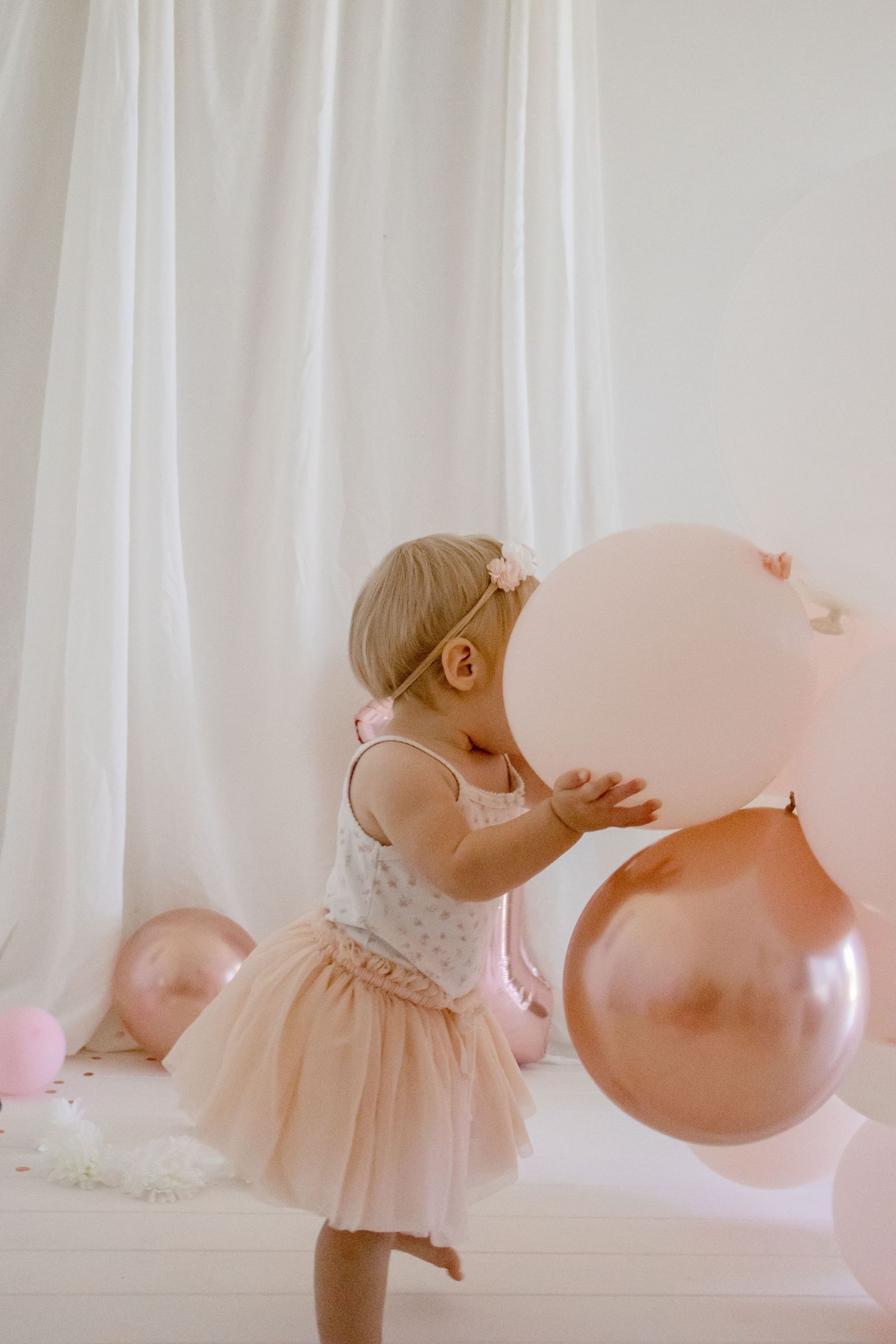 A toddler in a pink tulle dress plays with large pink and rose gold balloons.
