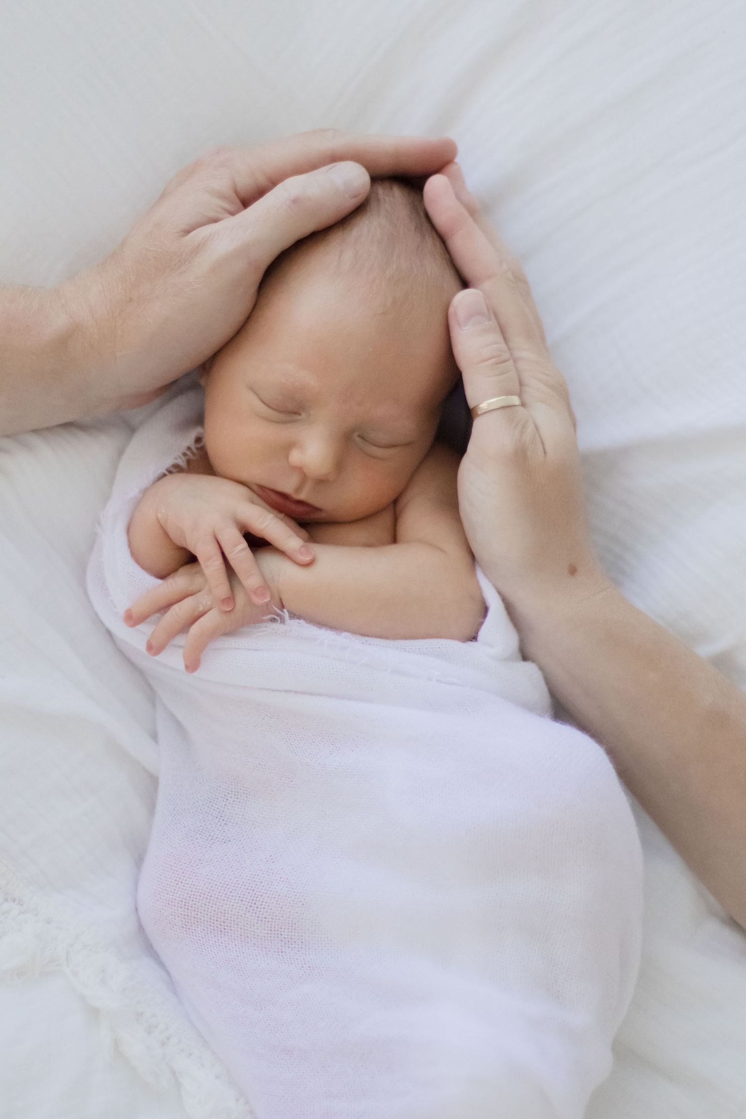 A sleeping newborn baby swaddled in white with gentle hands cradling their head on soft bedding.