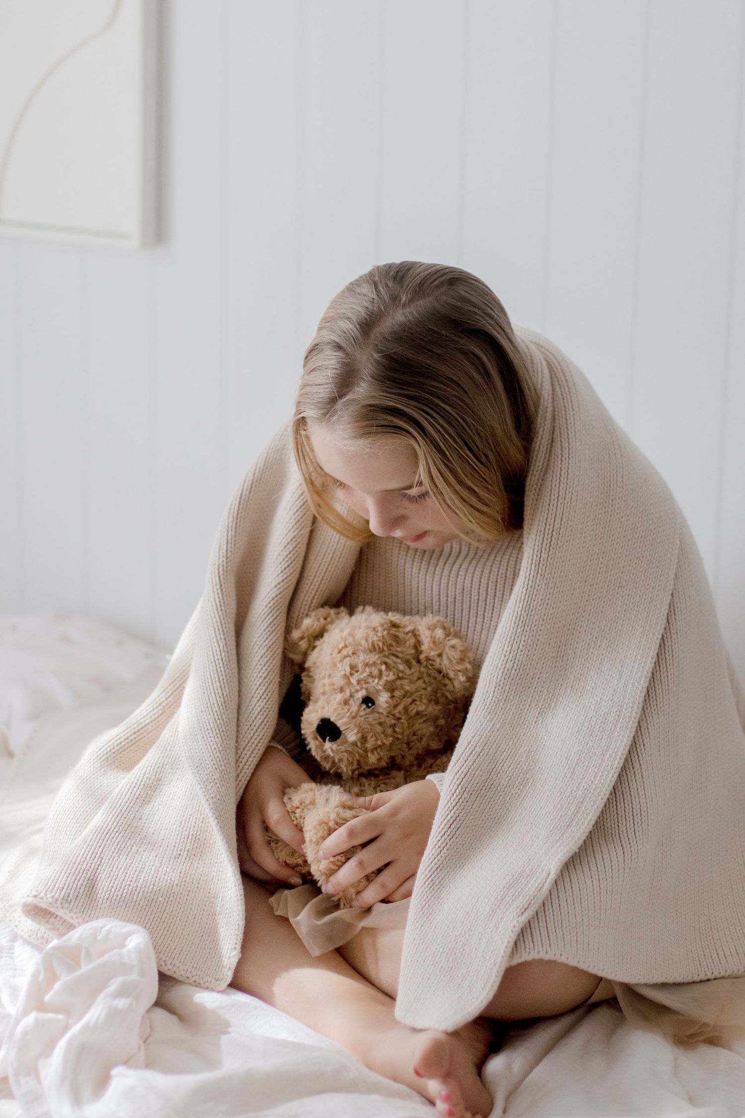 Person wrapped in cozy beige sweater and blanket cuddles with teddy bear on white bed.