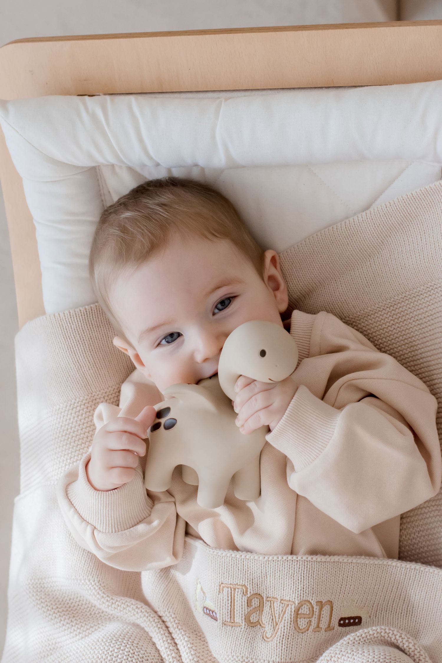 A baby wearing a cream-colored sweater lies on white bedding while playing and smiling.