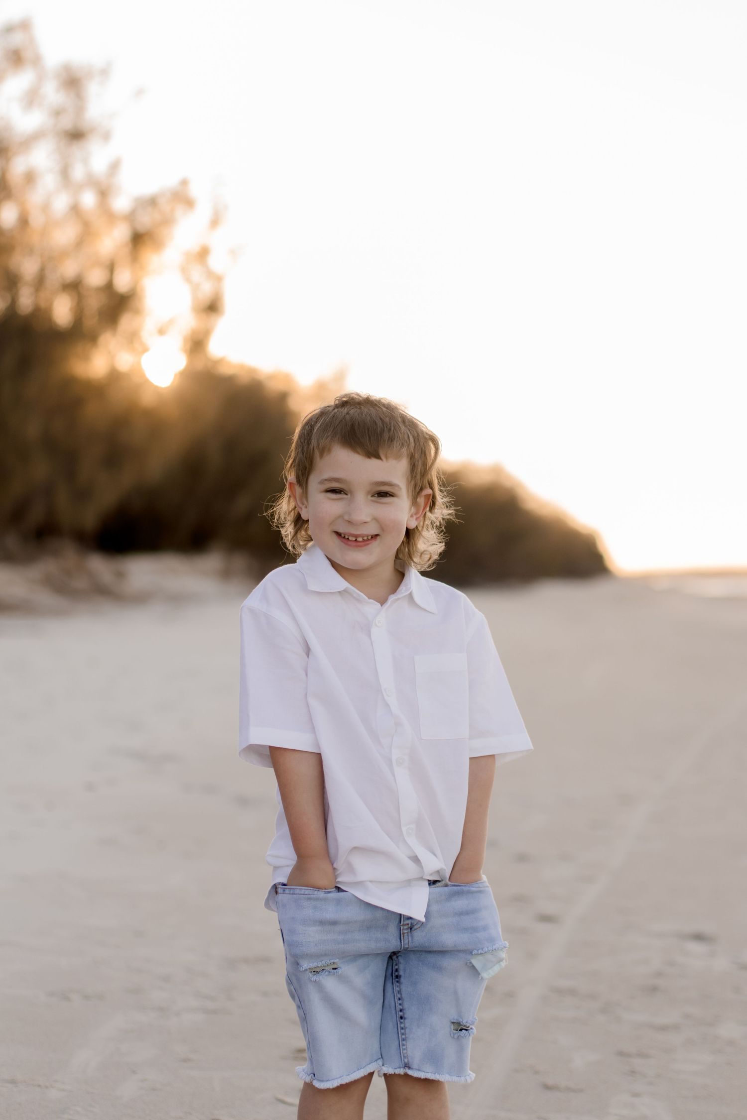 A young child in a white button-down shirt and denim shorts poses on a beach at sunset.