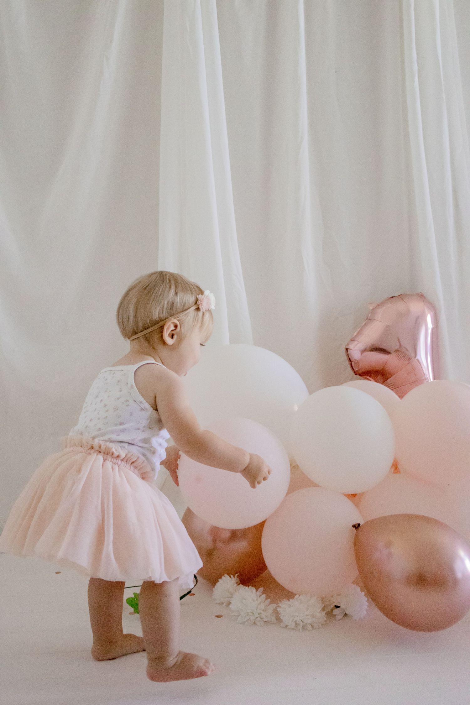 A small child in a tutu dress reaches for pink balloons against a white curtain backdrop.