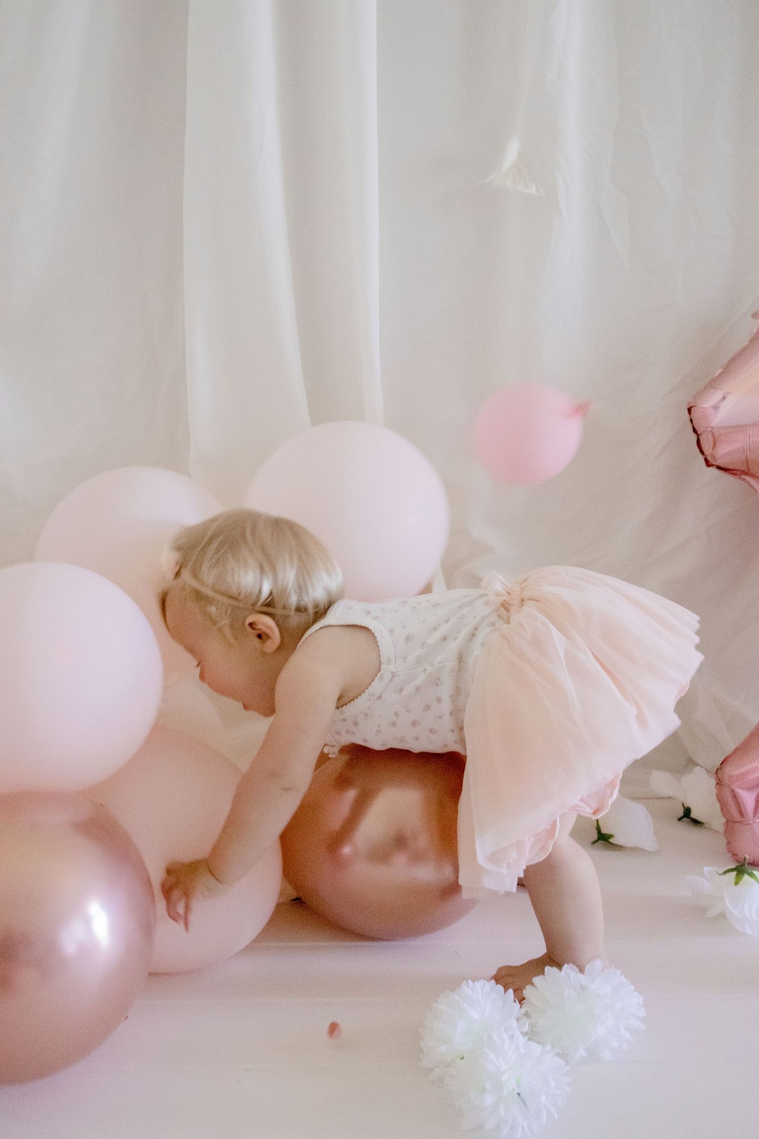 Baby in a pink tulle dress playing with pink and rose gold balloons against a white backdrop.