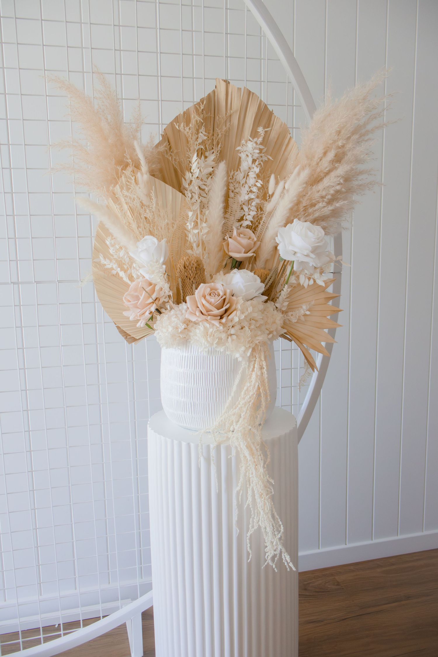 Boho dried floral arrangement with pampas grass, palm leaves, and blush roses in a white ribbed vase on a pedestal.