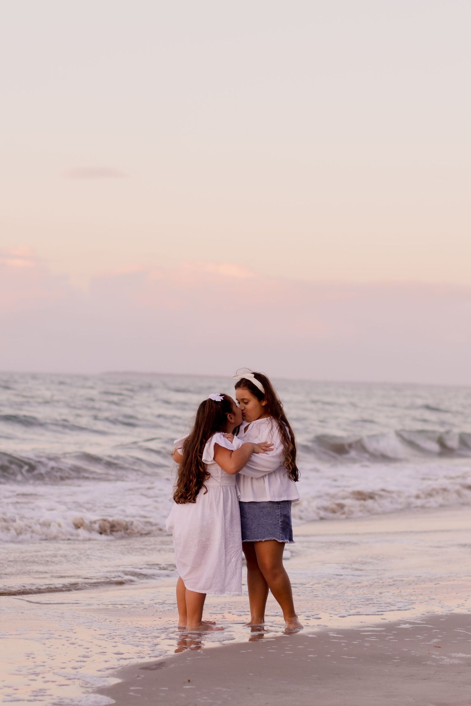 Two people in casual attire embrace while standing at the water's edge on a beach during sunset.