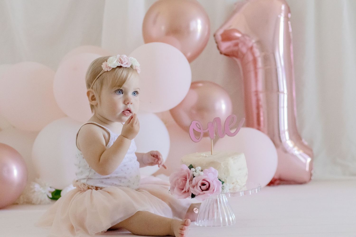 Baby in white dress sitting among pink party decorations for first birthday celebration.
