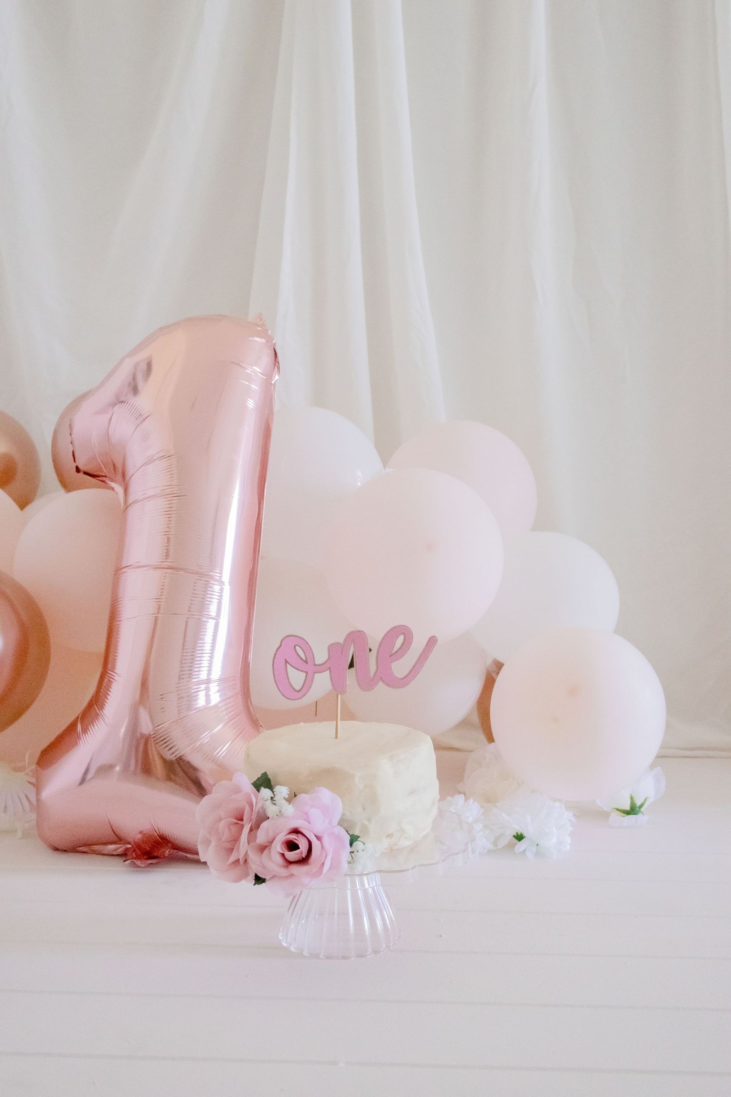 Rose gold number one balloon with white cake and pink roses on glass pedestal.