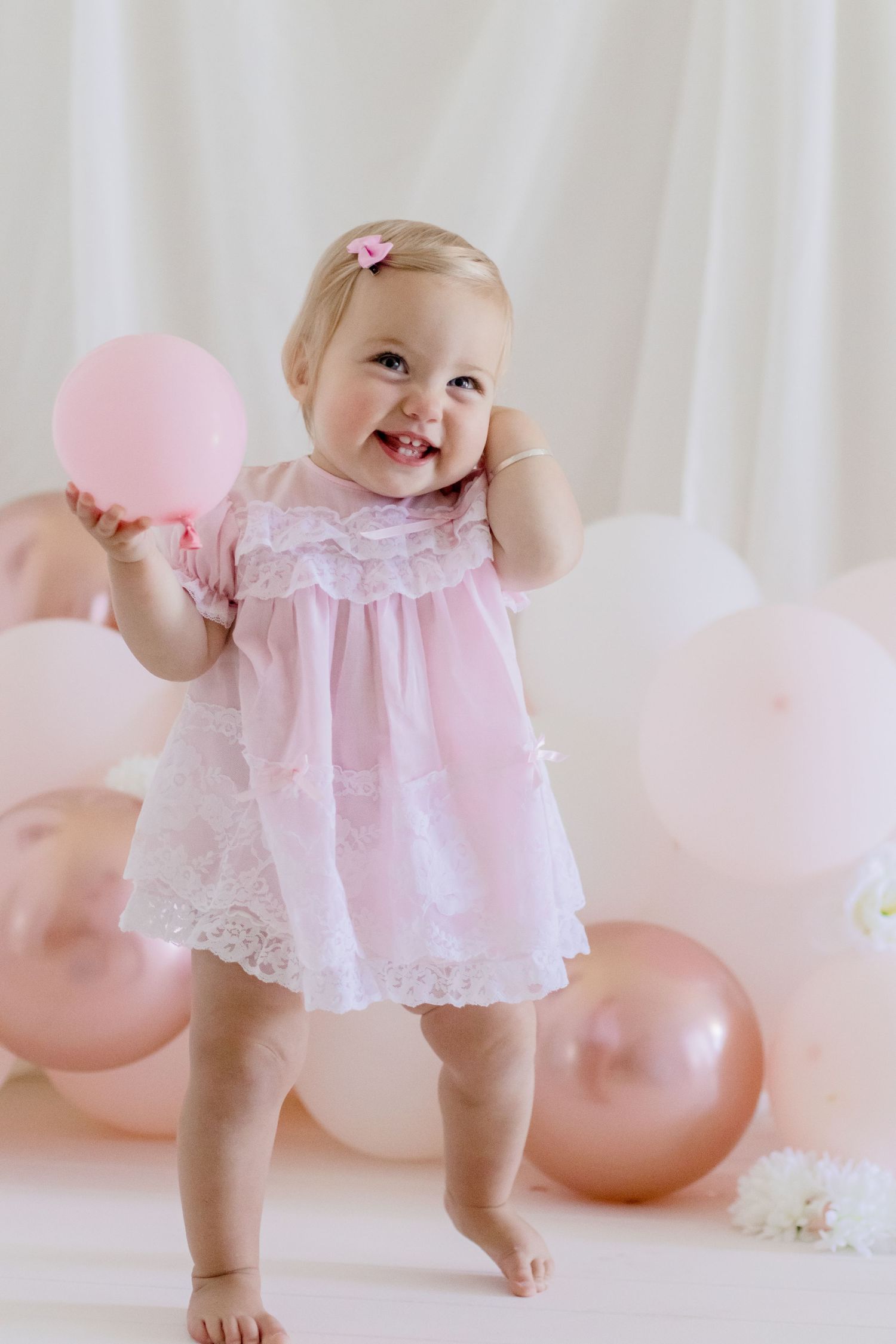 Happy toddler in pink dress holding balloon against white backdrop with rose gold decorations.