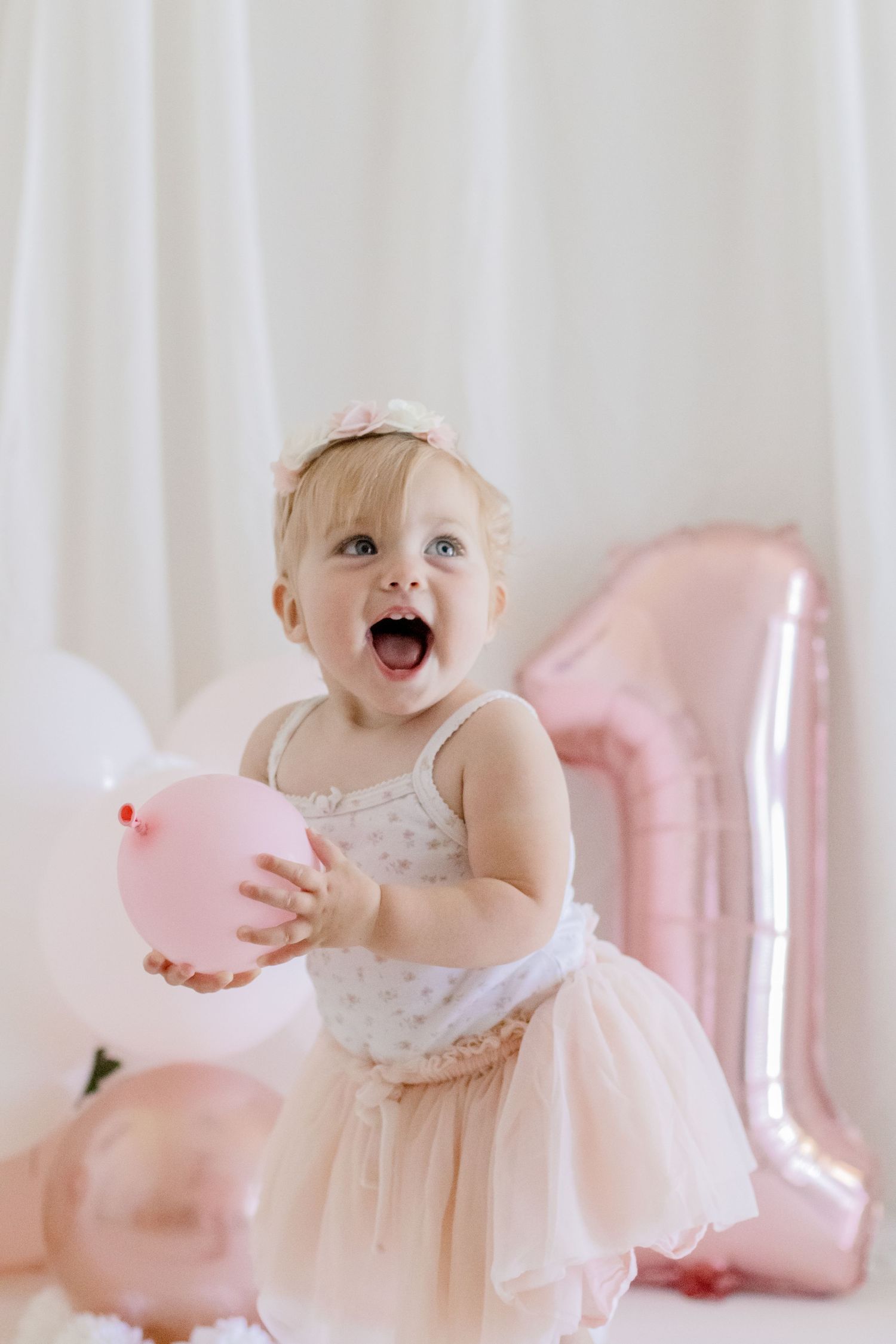 Excited toddler in pink tutu dress playing with balloons against white backdrop.
