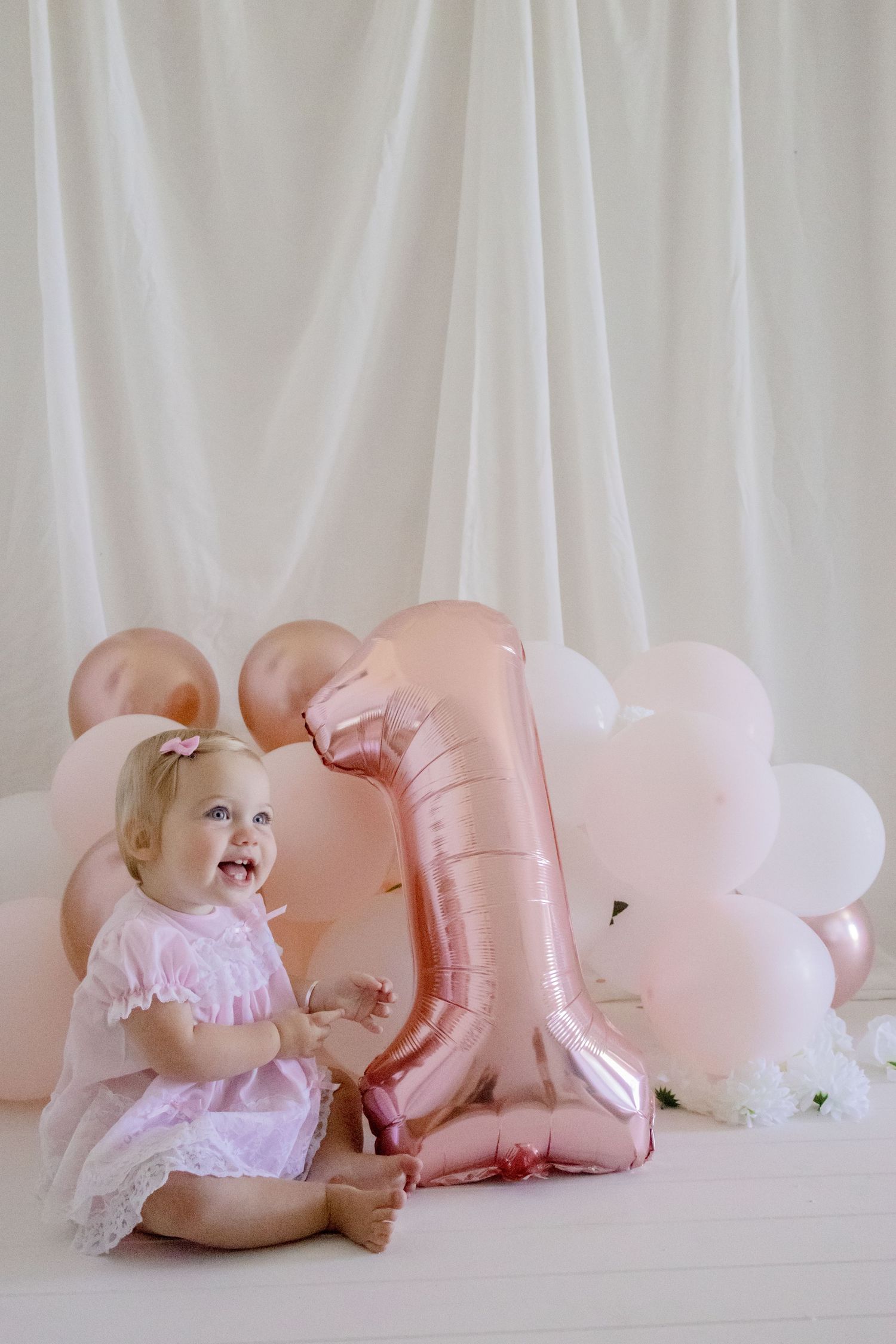 Laughing baby in pink dress sitting beside rose gold number one balloon against white curtain.