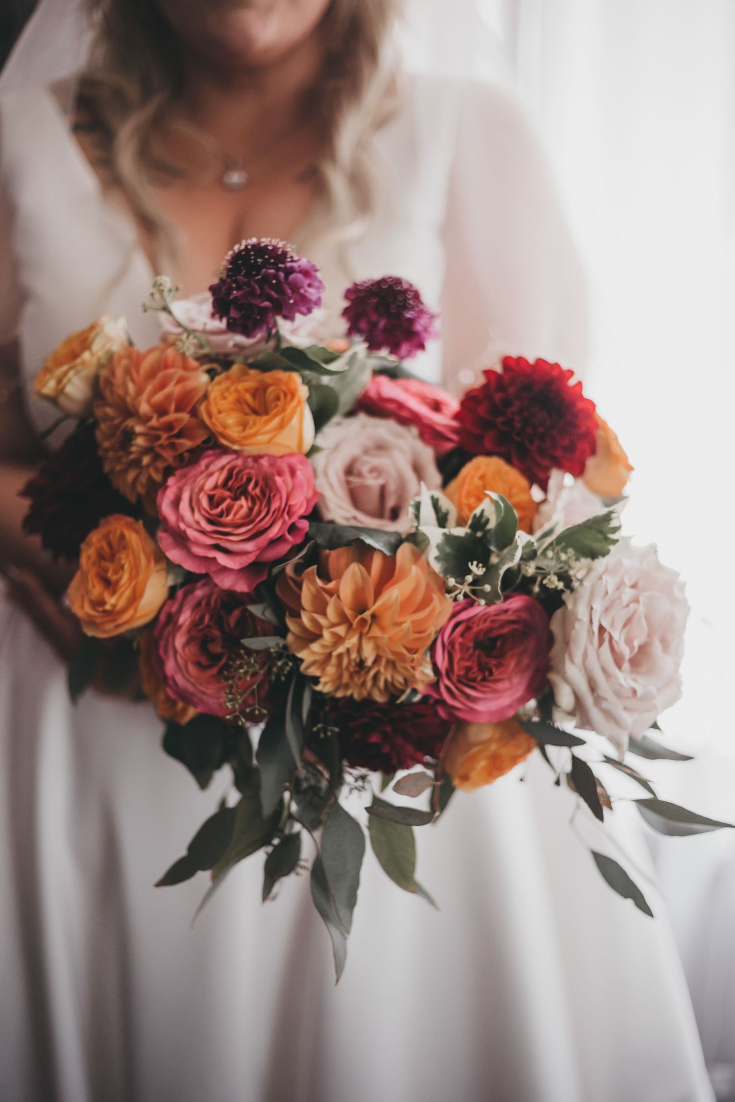Vibrant autumn bouquet of orange, pink and burgundy roses and dahlias with eucalyptus held against white fabric.