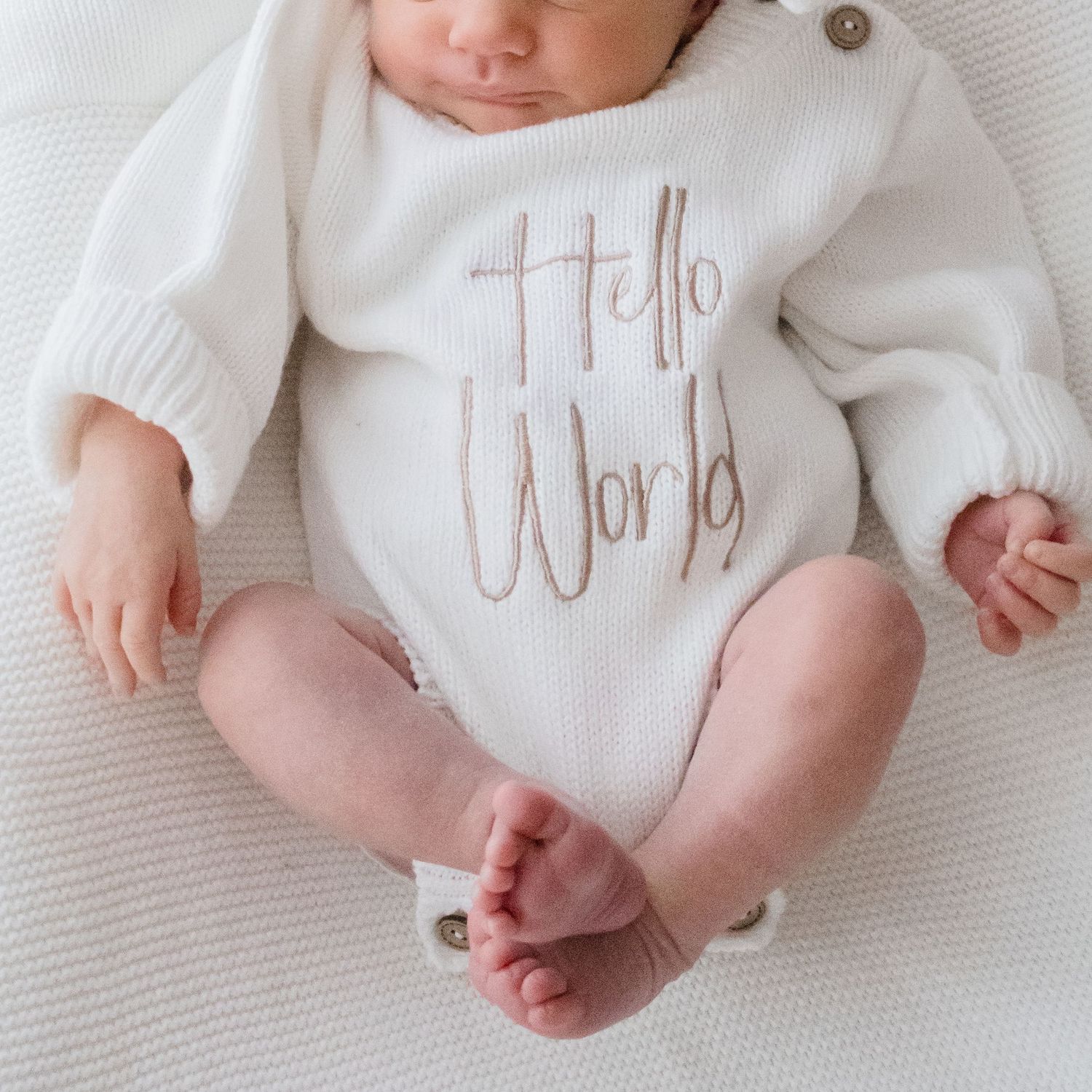 Baby wearing white knit sweater with 'Hello World' text laying on white textured surface.