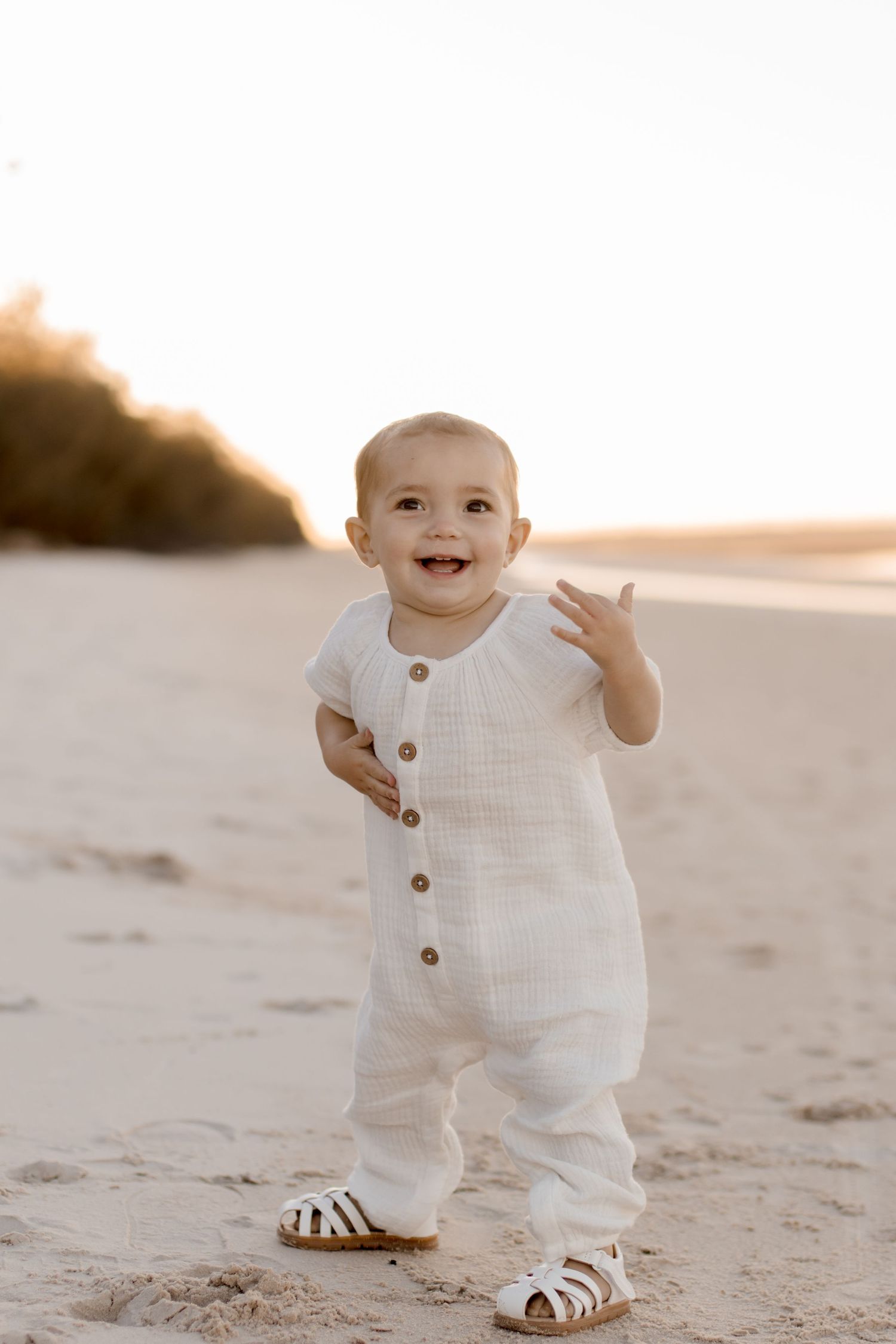 Toddler wearing white linen romper and sandals waves while standing on sandy beach at sunset.
