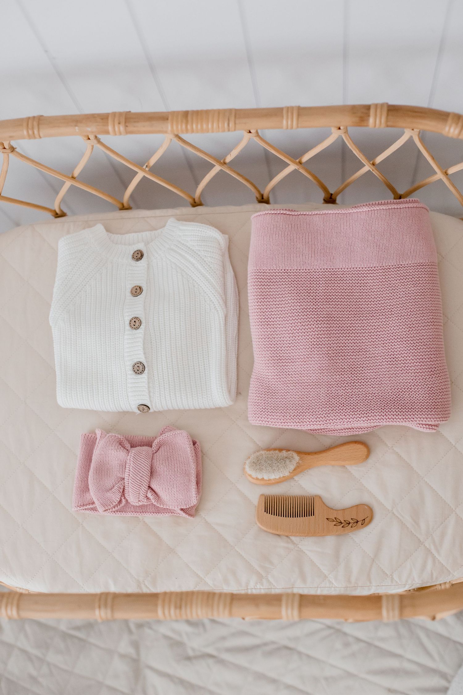 Flatlay of baby items including white knit cardigan, pink blanket, bow headband, and wooden brush on rattan changing table.