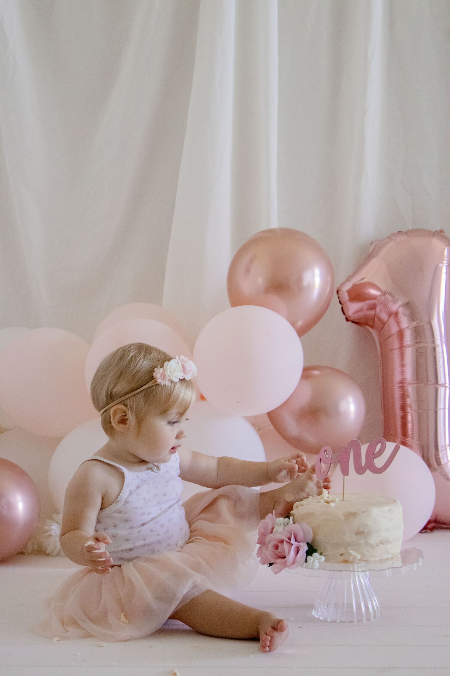 A cake smash photo session with pink balloons, roses, and white cake on a white backdrop.