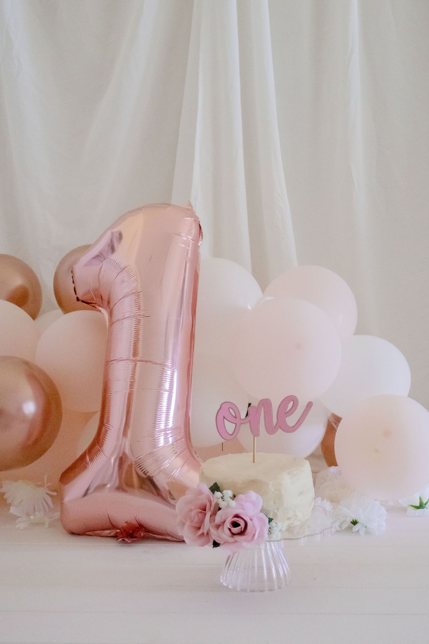 First birthday cake setup with rose gold number one balloon and pink roses against white backdrop.