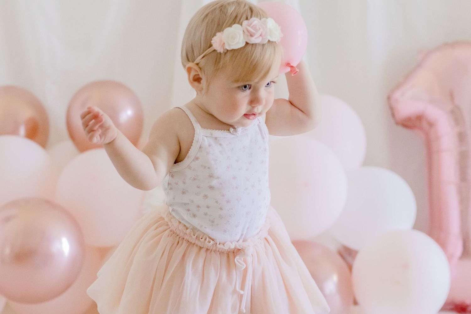 Small toddler in a white and pink tulle dress playing with pink and rose gold balloons against a white backdrop.