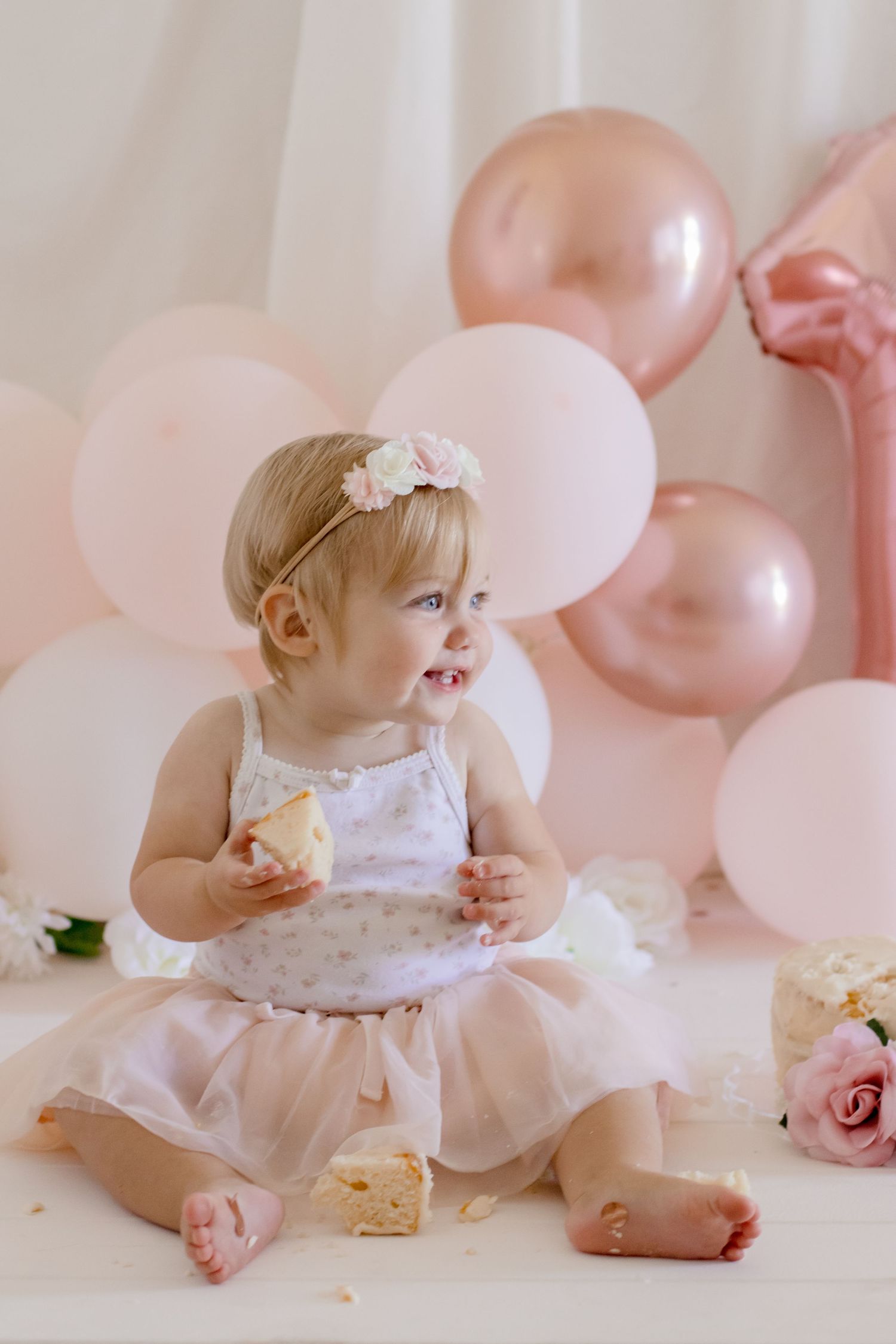 Baby in tutu dress sitting on floor with pink and rose gold balloon decorations.