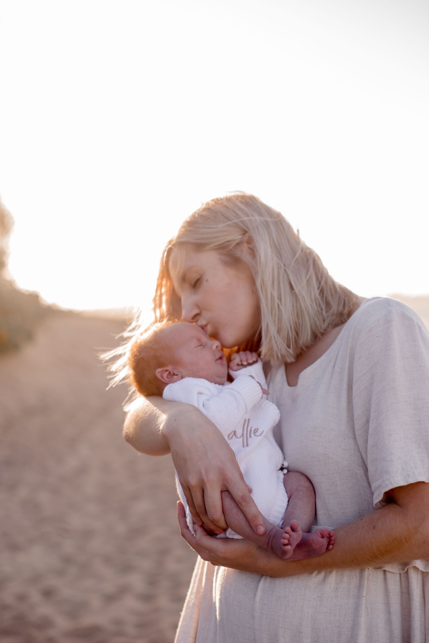Intimate moment captured at sunset as parent holds newborn in white outfit on sandy beach.