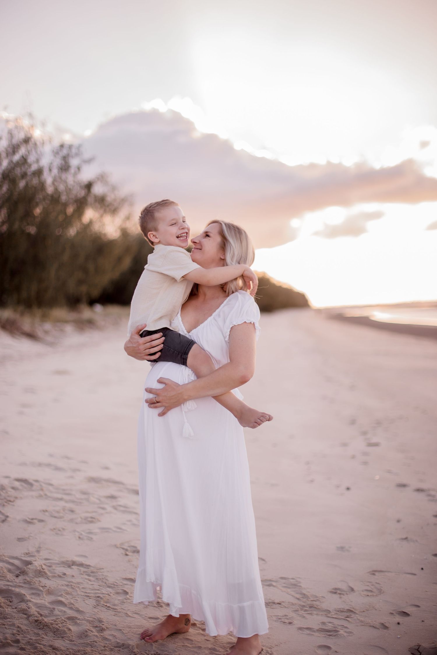 Mother in flowing white dress holds and embraces young child on beach during sunset.
