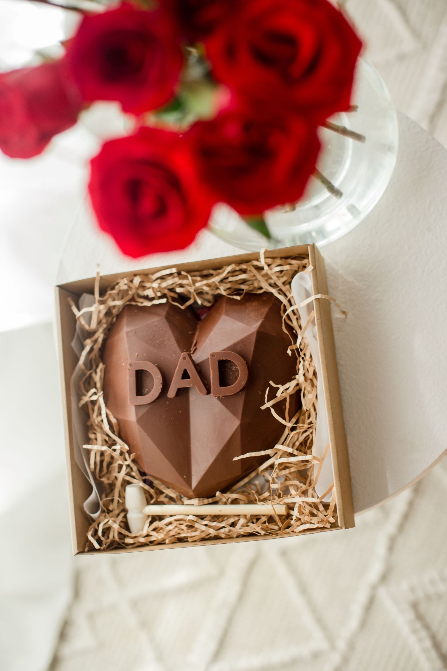 Chocolate heart with 'DAD' text in wooden gift box with red roses in background.