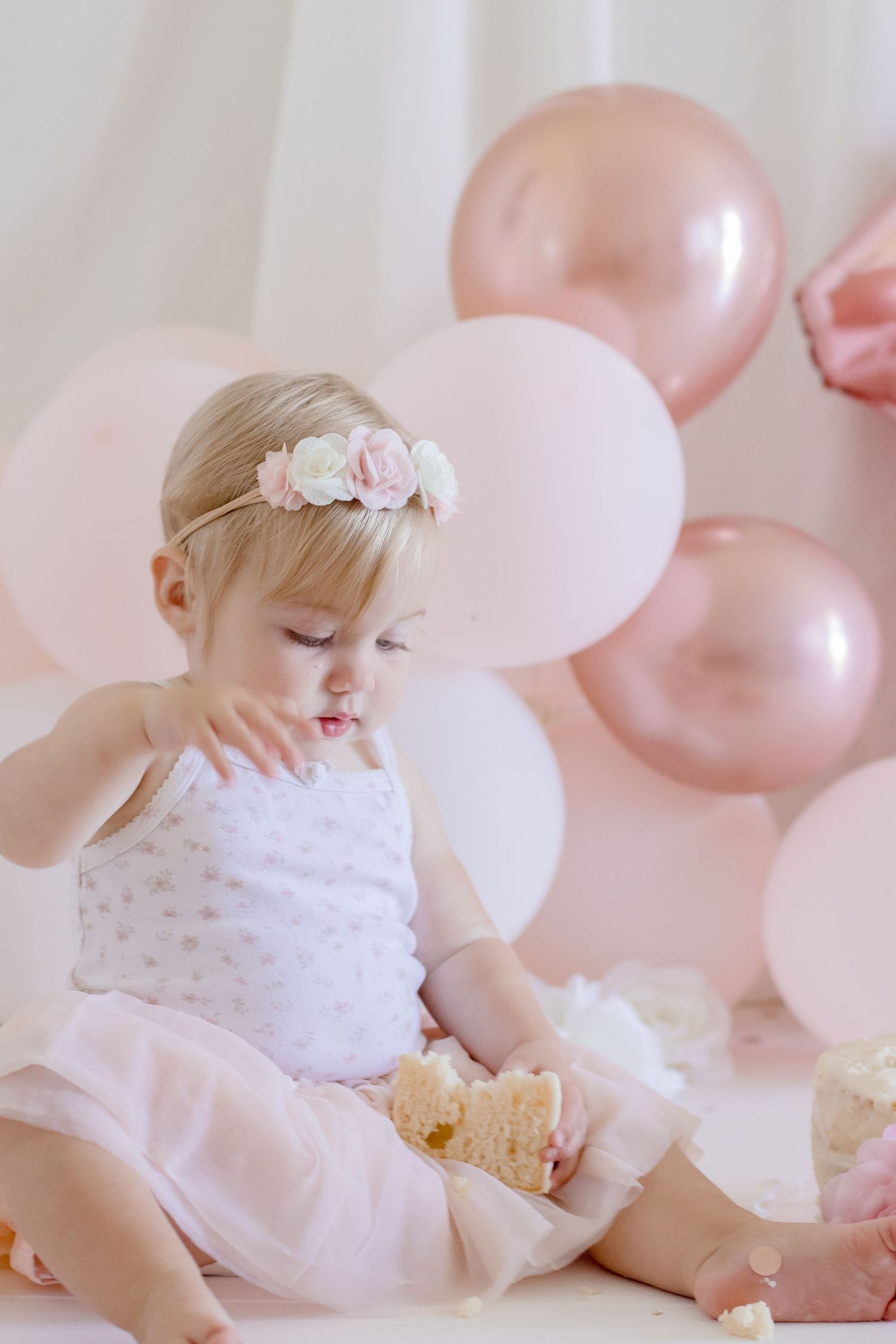 Baby in white dress with floral crown eating snack against pink balloon backdrop.