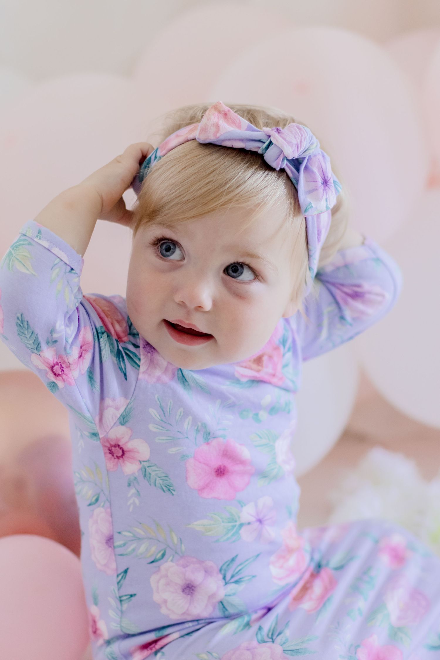 Baby wearing floral outfit touches matching headband during photo session.