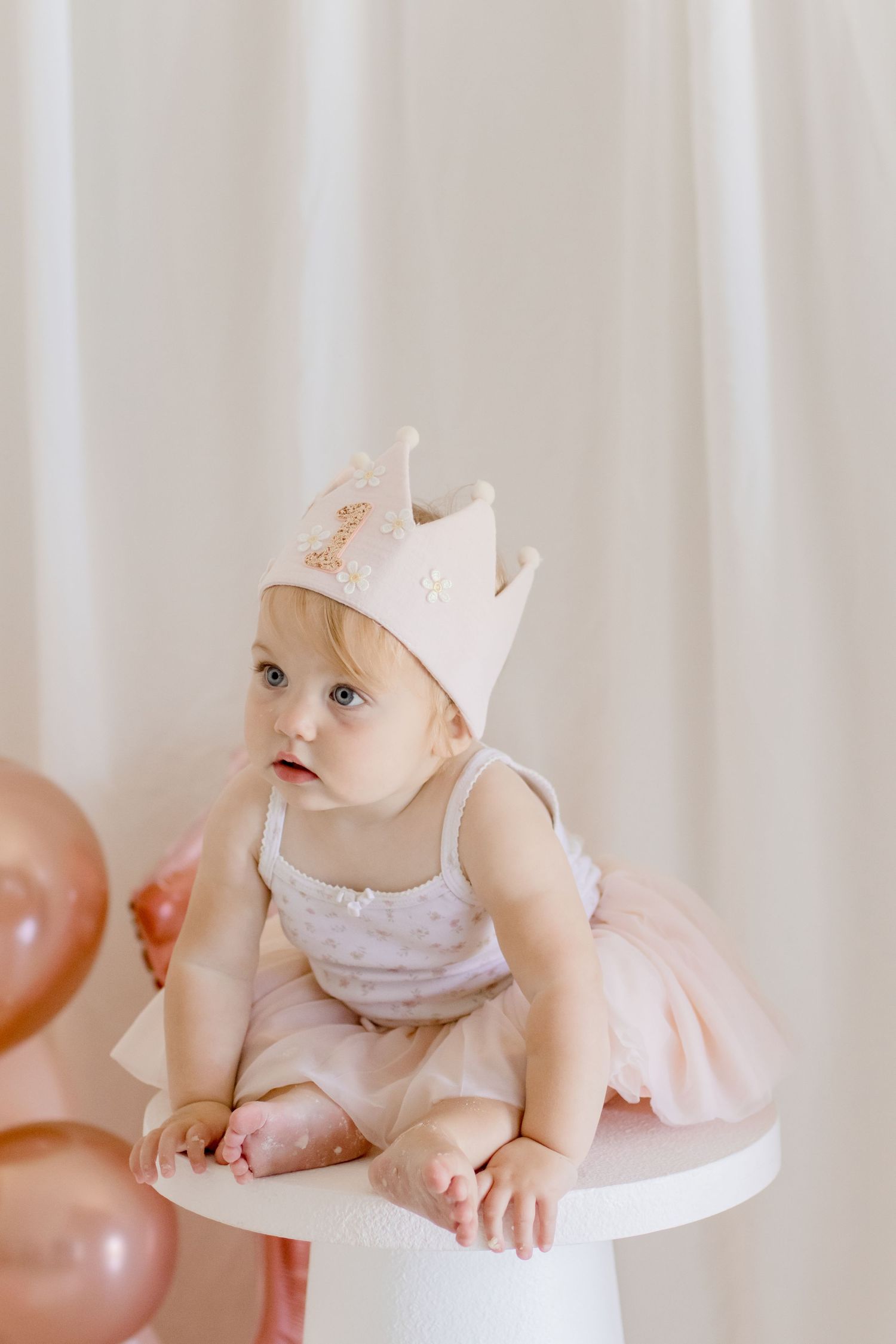Baby in a light pink tutu and crown sits on white pedestal with pink balloons.