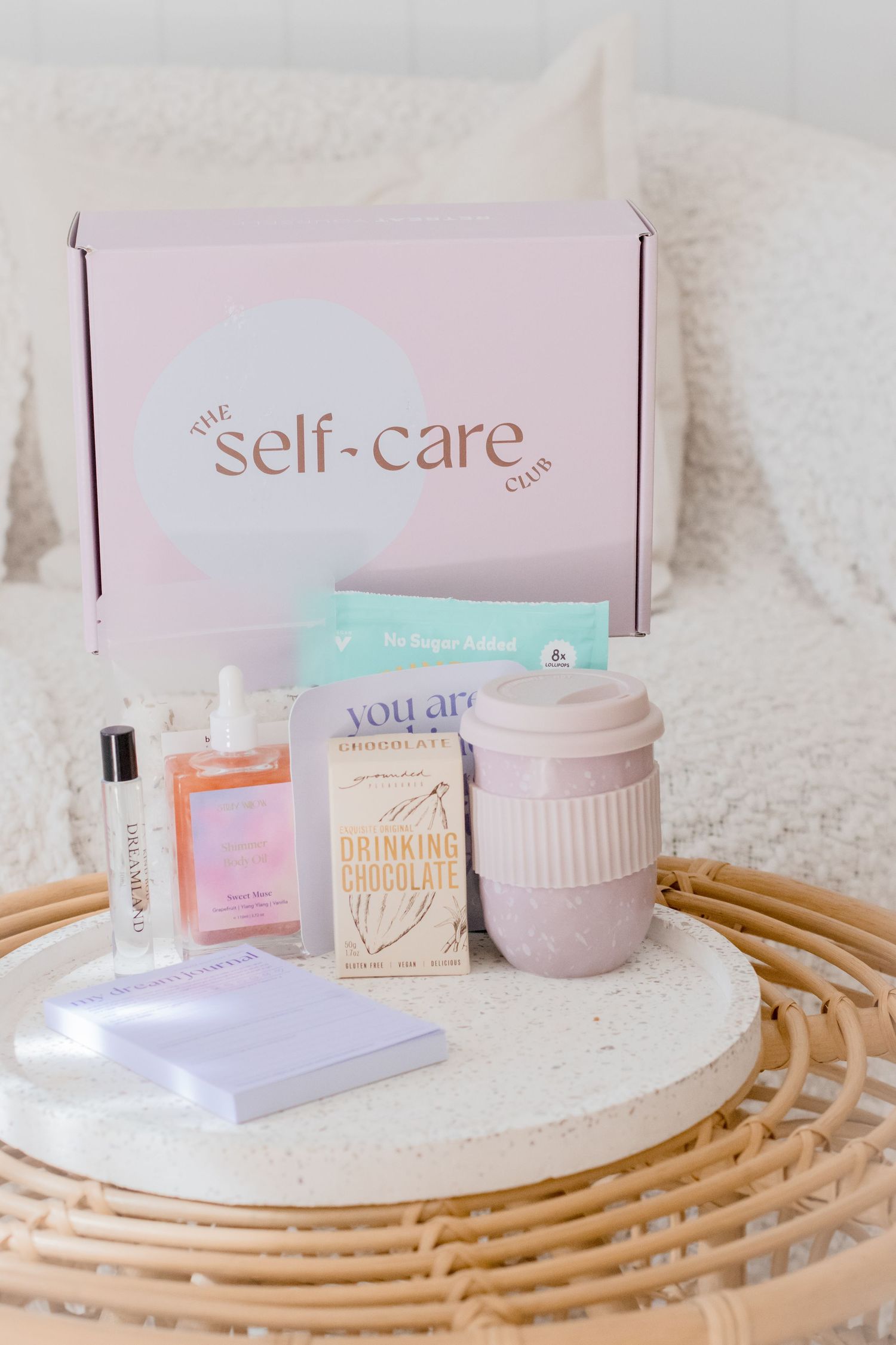 A self-care subscription box with pink packaging, a reusable cup, and wellness products displayed on a rattan surface.