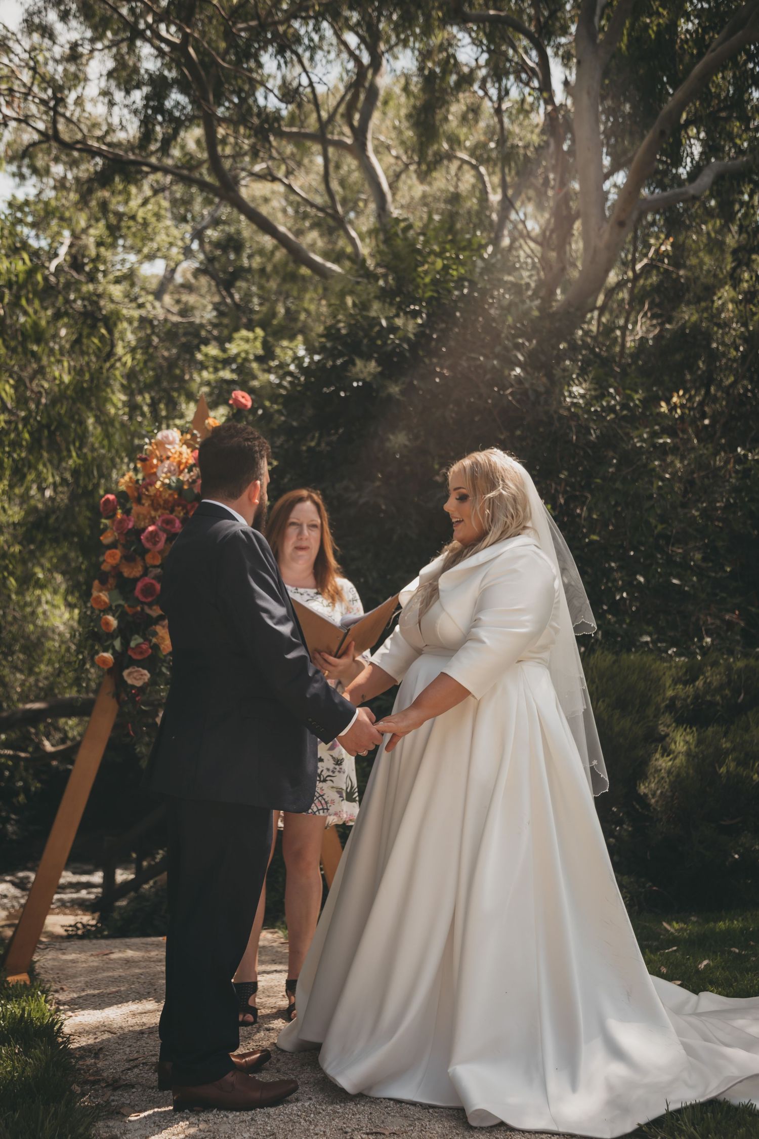 A wedding ceremony takes place outdoors in a natural setting with a beautiful floral arch and greenery backdrop.