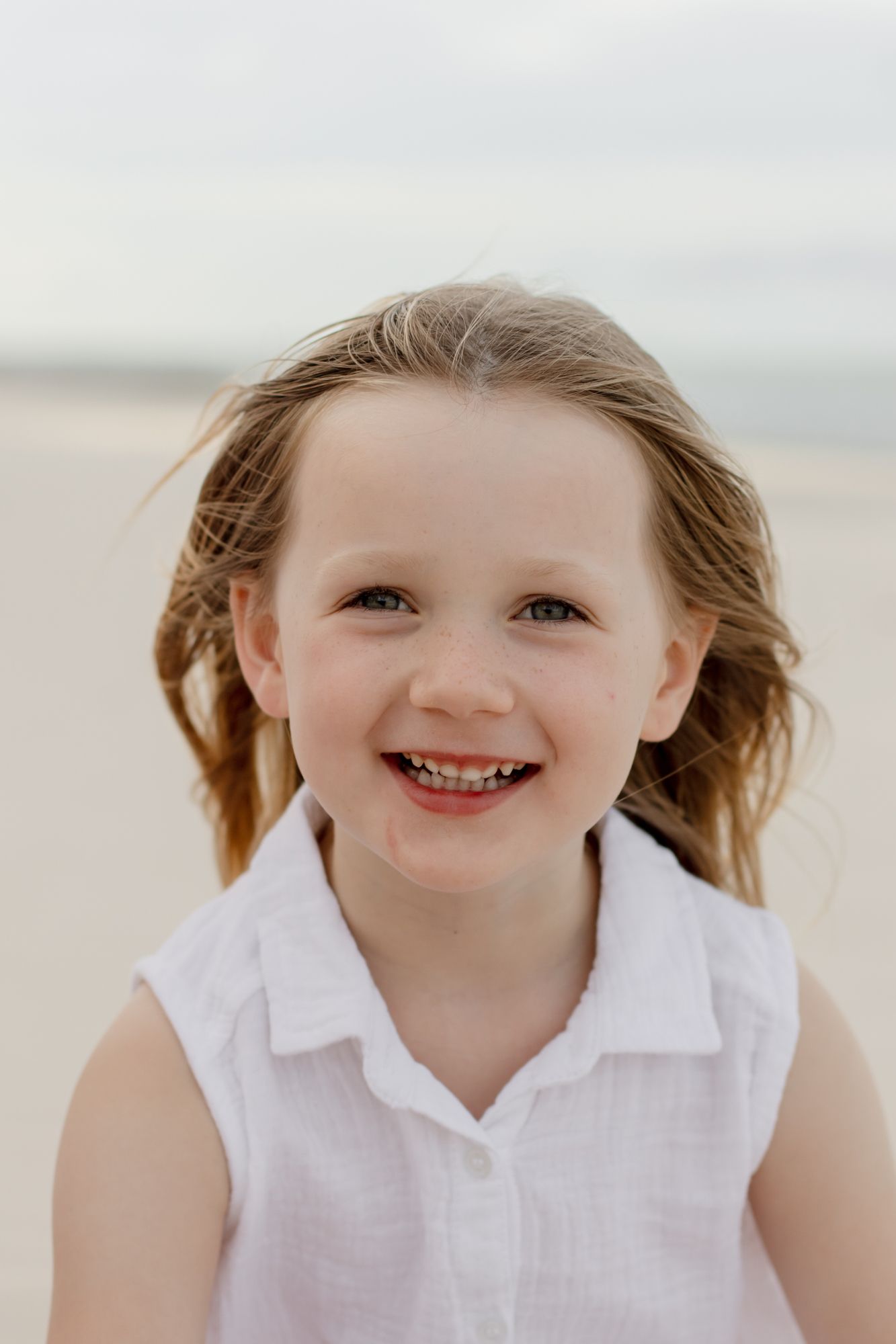A smiling child with windswept hair poses on a beach.