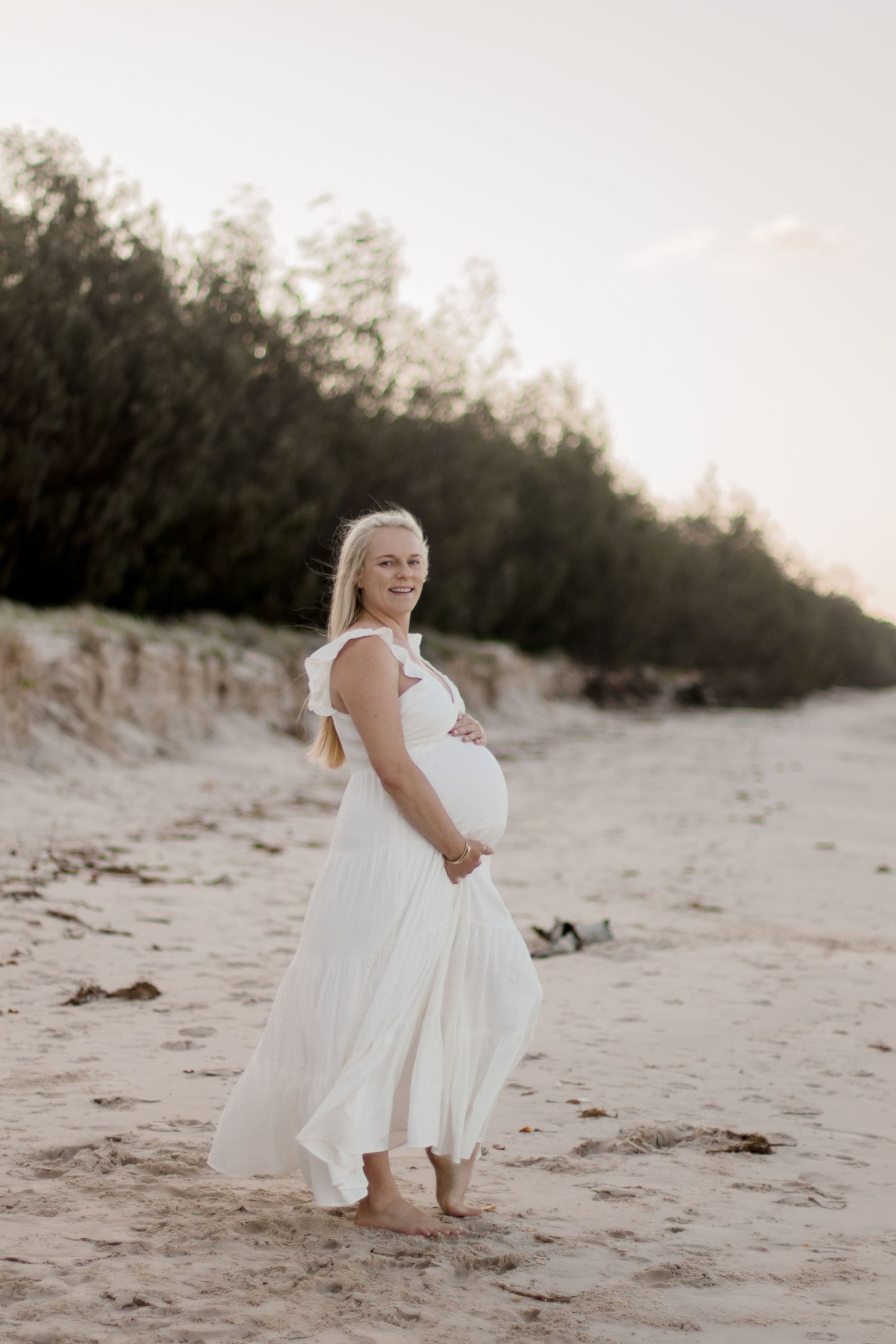 An expectant mother in a flowing white dress poses on a beach during golden hour.
