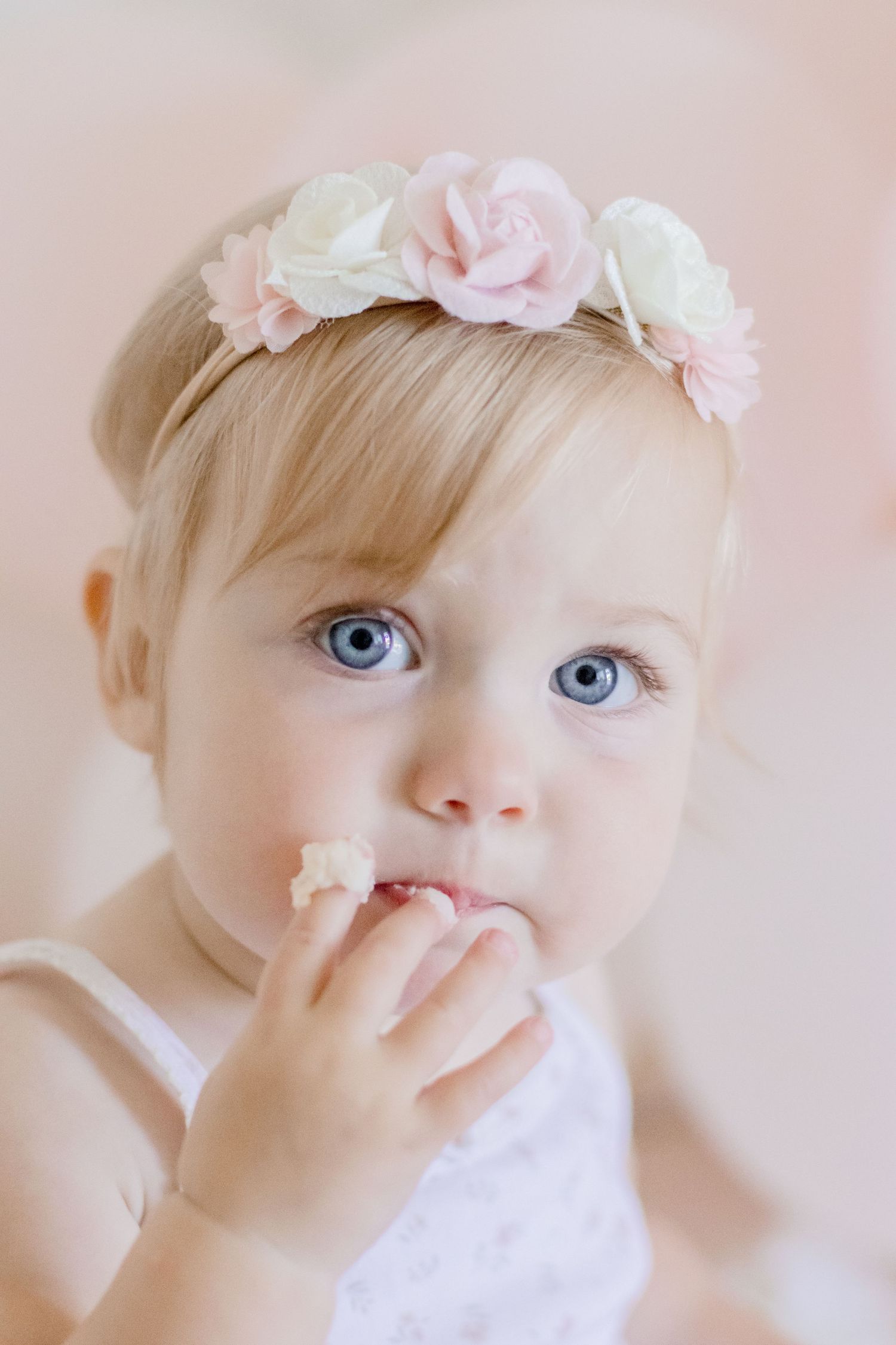 Close-up portrait of baby with floral crown and blue eyes touching their lips.