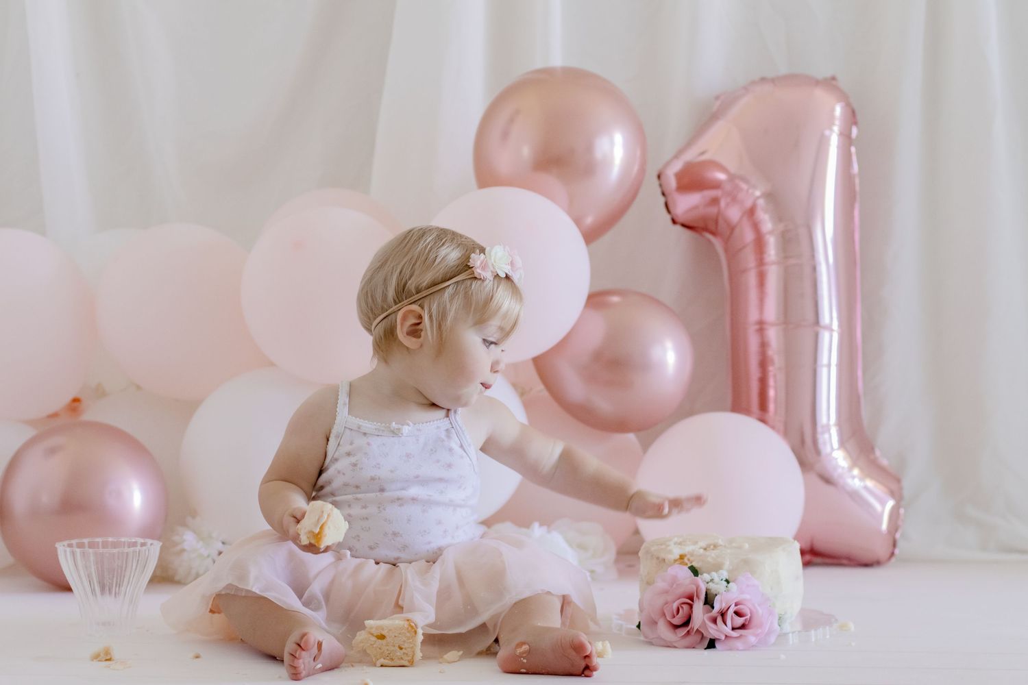 Baby in white tulle dress with number 1 balloon and pink party decorations.