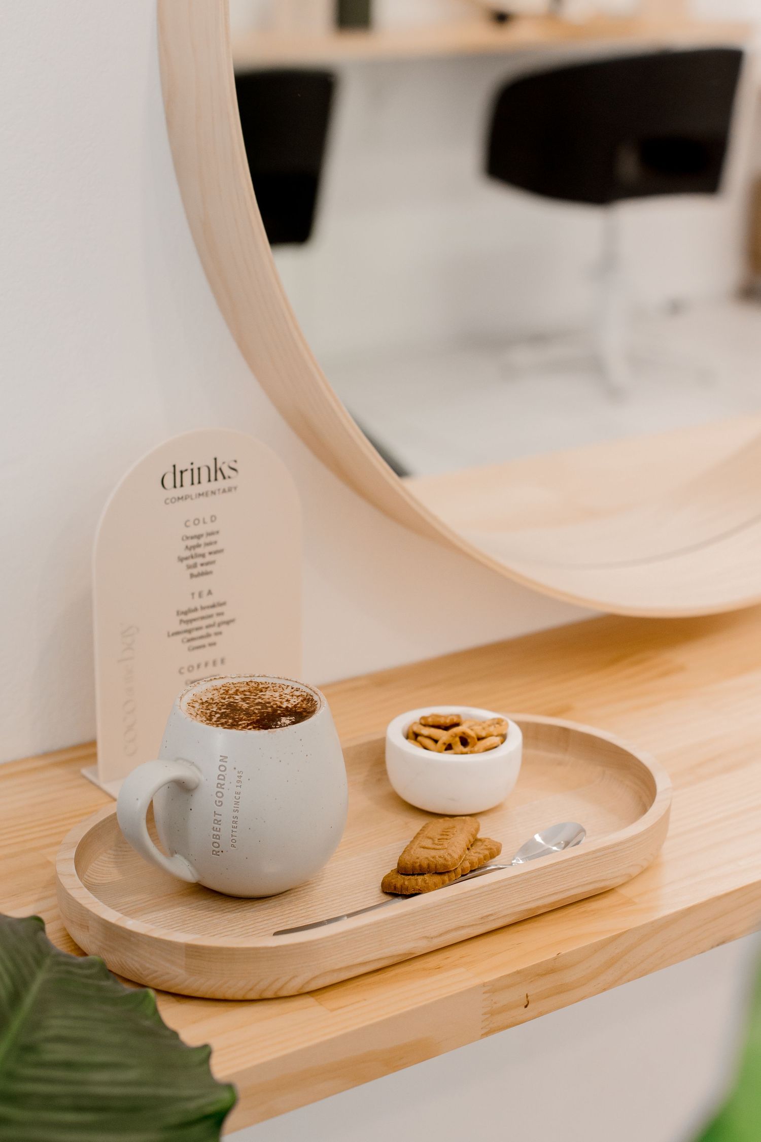 A cozy coffee setup with a white mug, snack bowl, and drink menu on a wooden tray near a circular mirror.