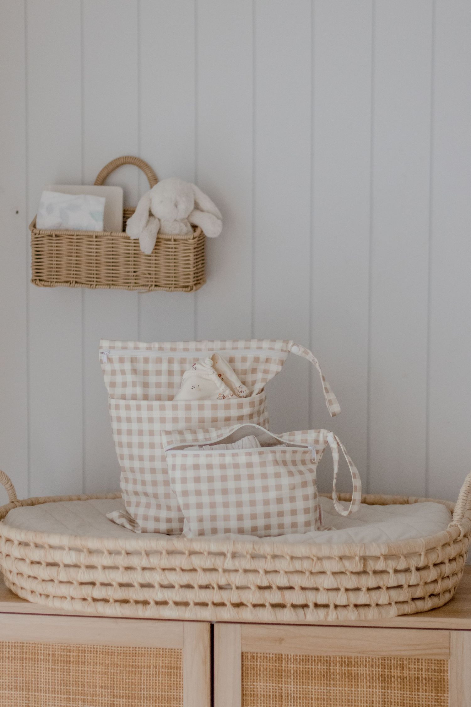 Neutral nursery storage with woven basket and gingham bags on wooden furniture.
