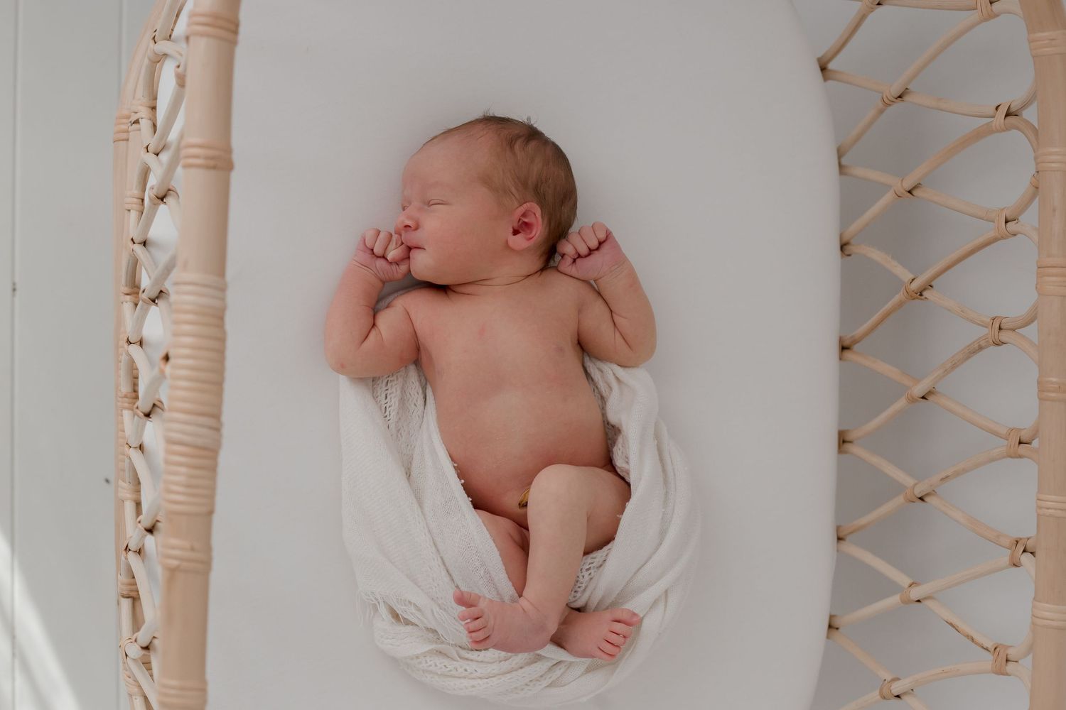 A newborn baby sleeps peacefully in a white basket with woven decor elements visible in the background.