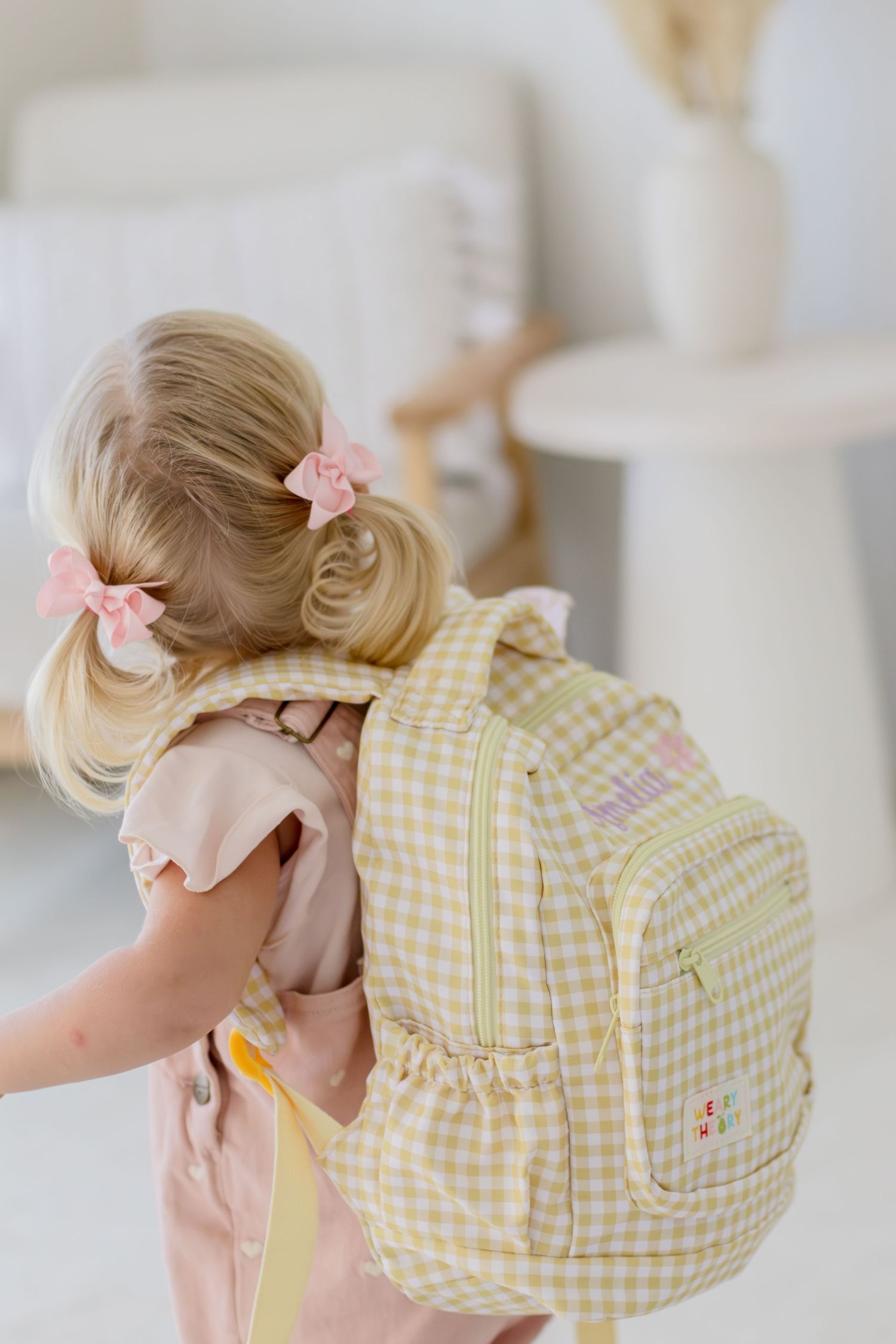 Toddler girl with blonde pigtails wearing a yellow gingham backpack with floral embroidery in a bright room.