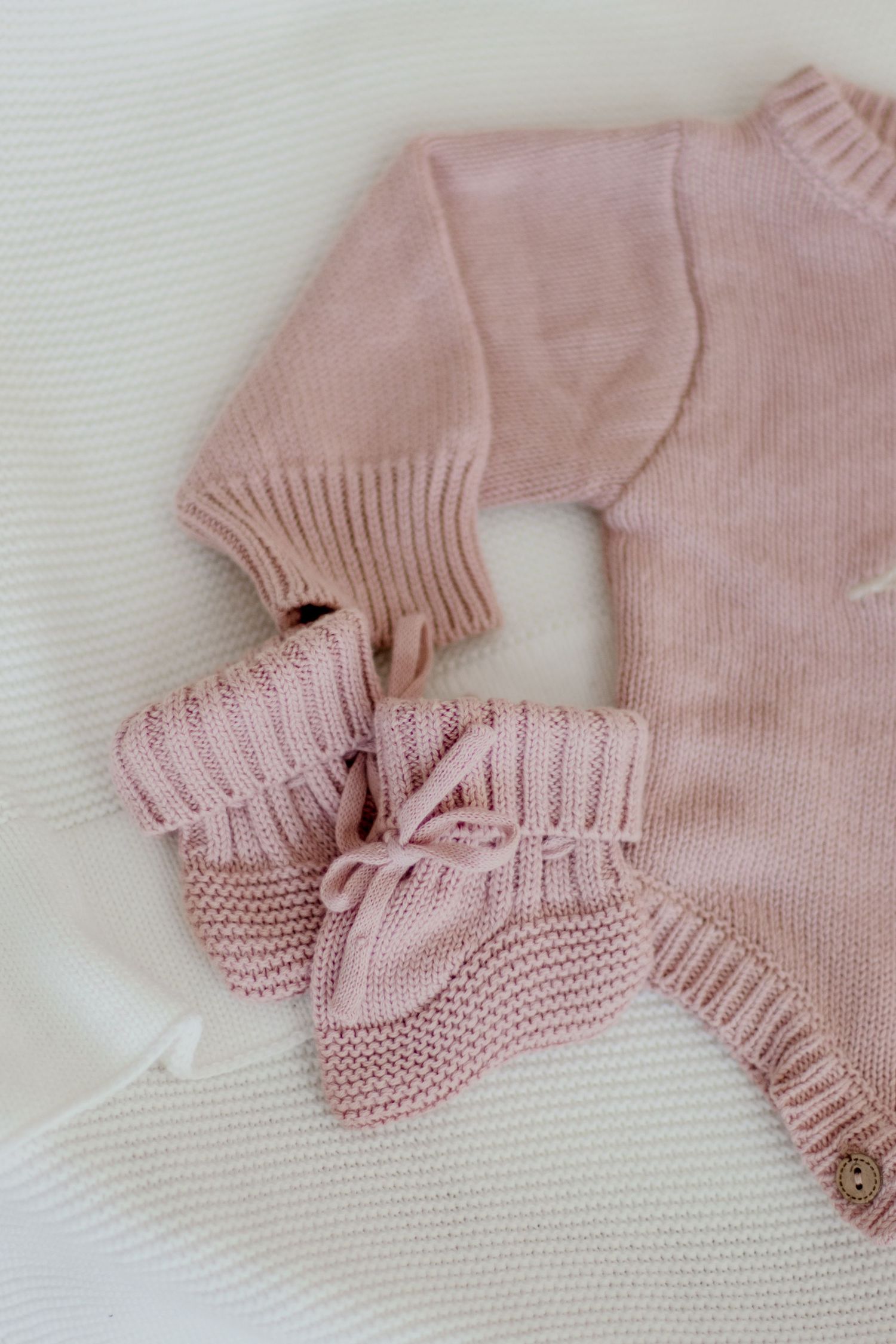 Soft pink knitted baby booties and sweater lying on white textured fabric in minimalist style.