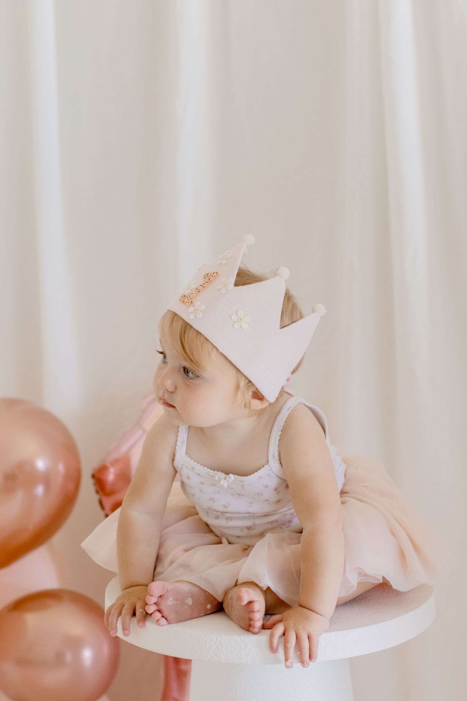 Baby in a tulle dress and crown looks thoughtfully at balloons while sitting on white pedestal.