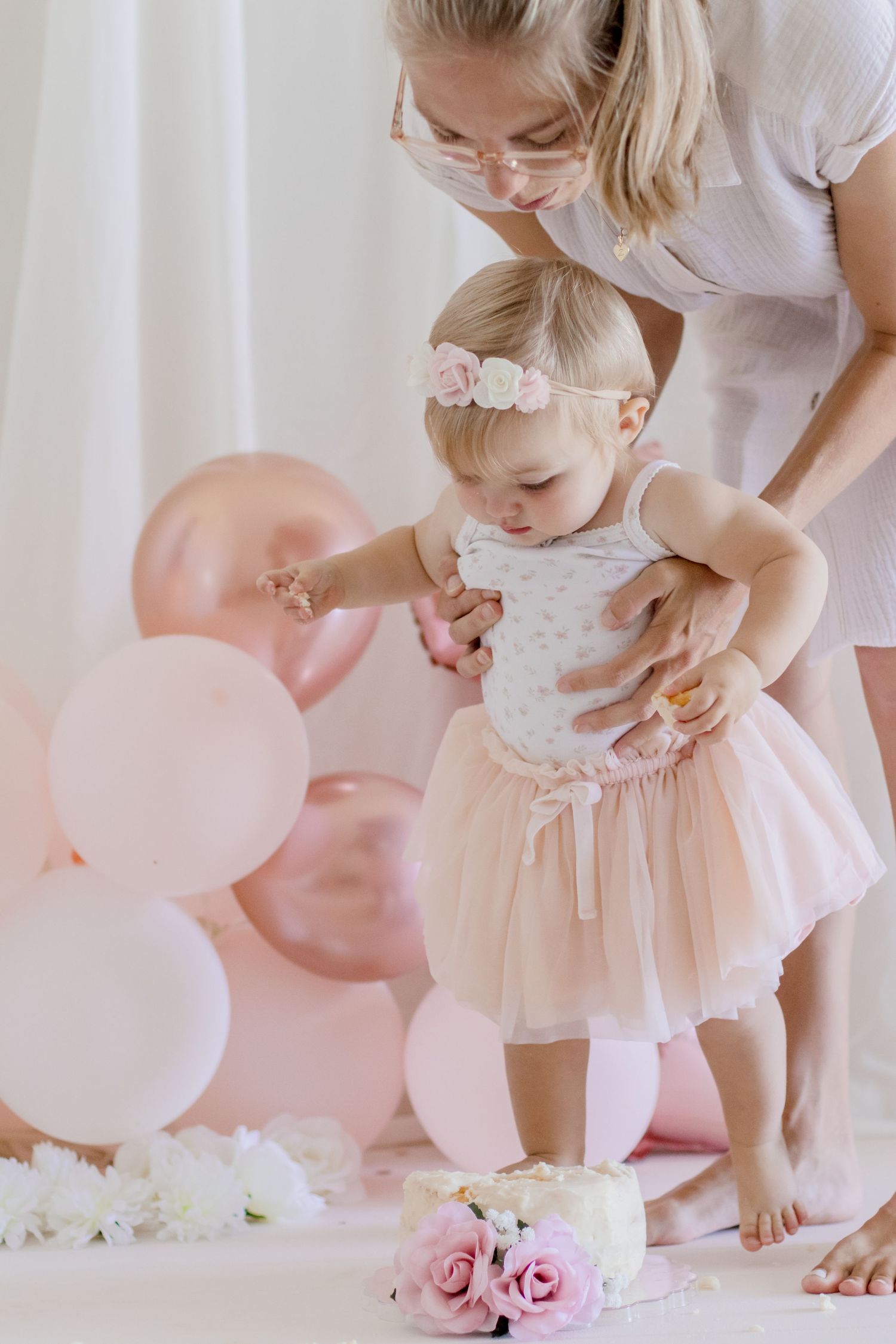 Baby taking first steps in tulle dress with pink balloon party decorations.