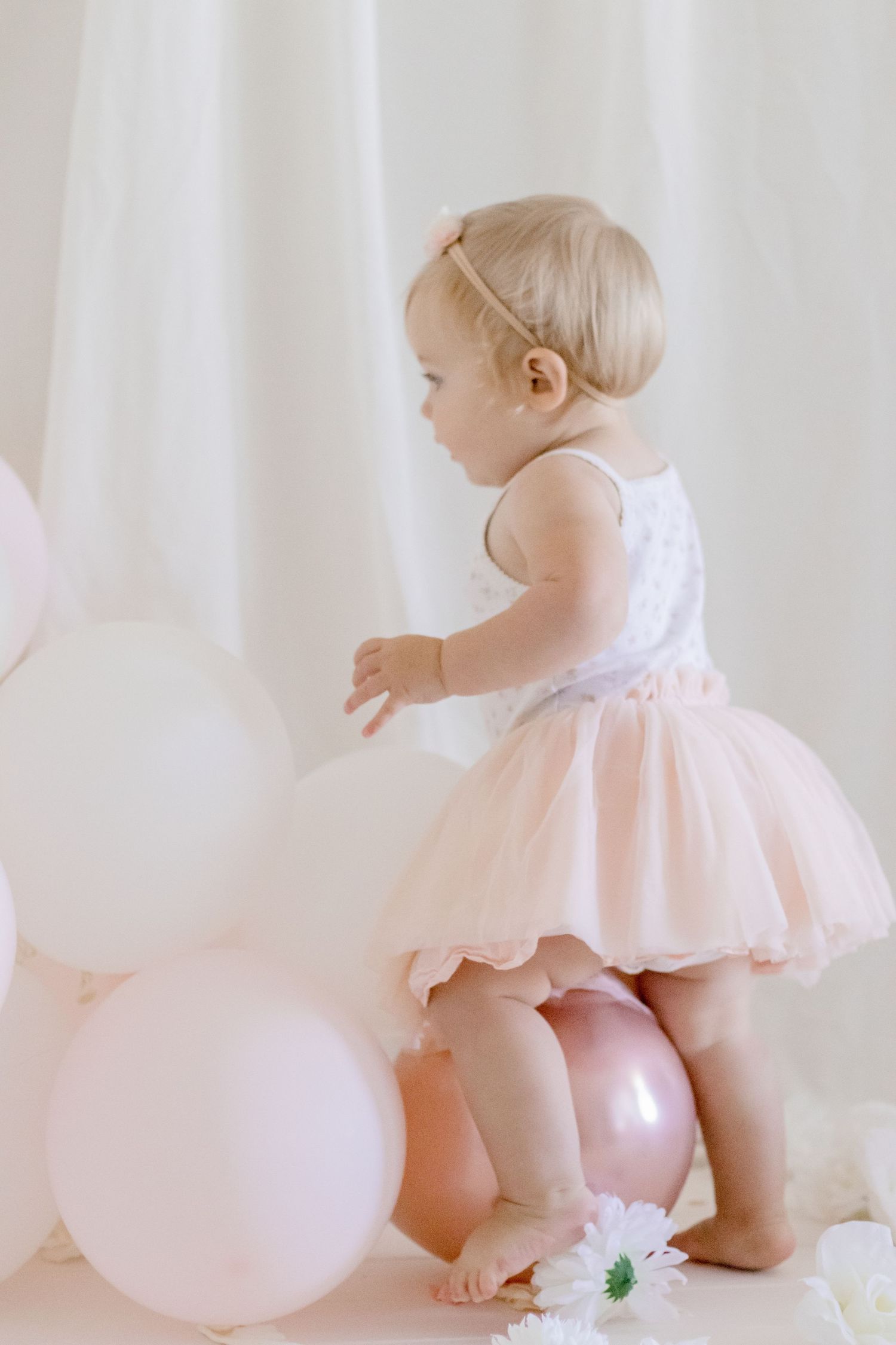 Small child in a ballerina outfit reaching for pink balloons in a dreamy photoshoot setting.