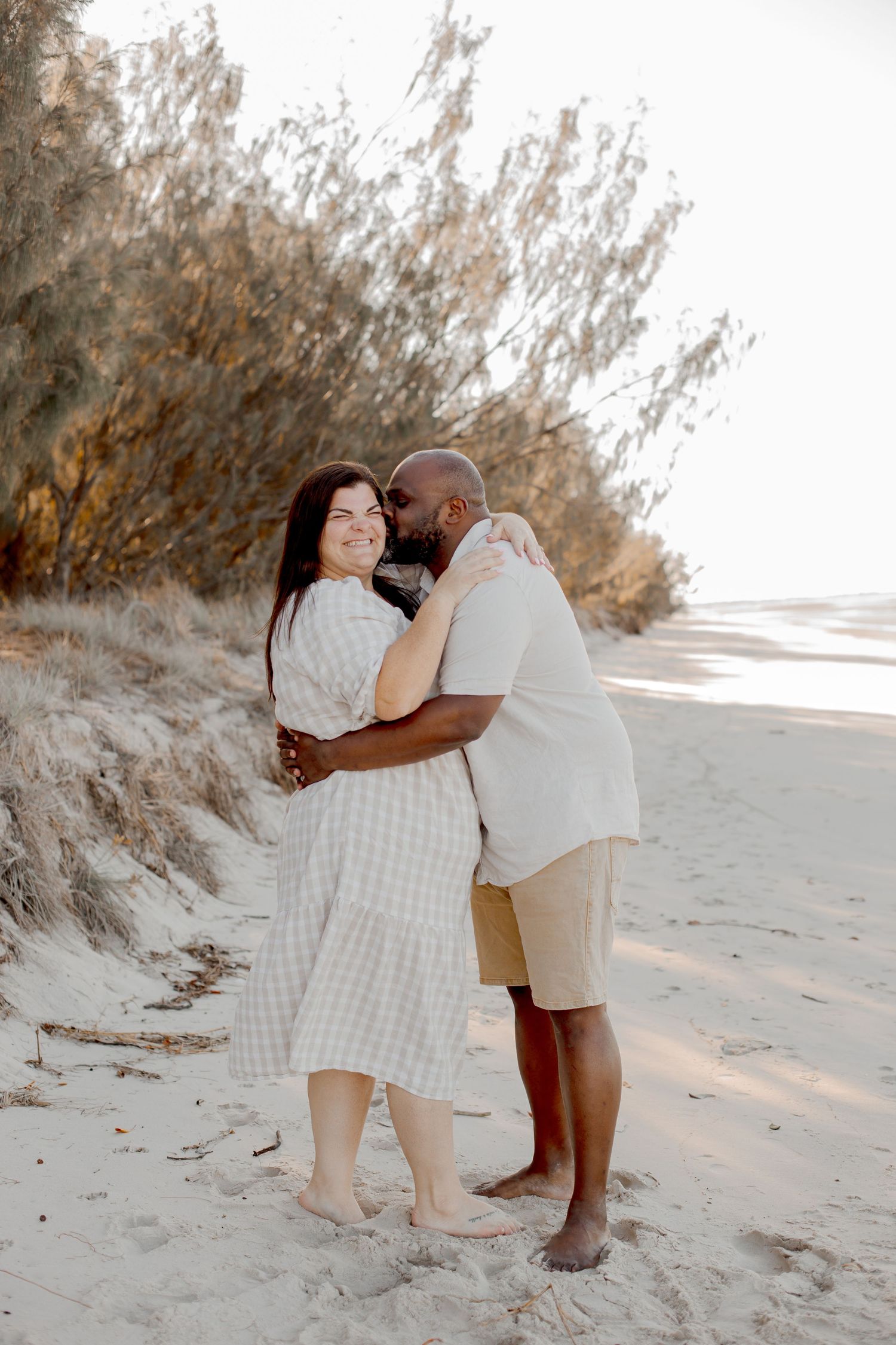 Couple embracing on a beach with sand dunes in the background at sunset.