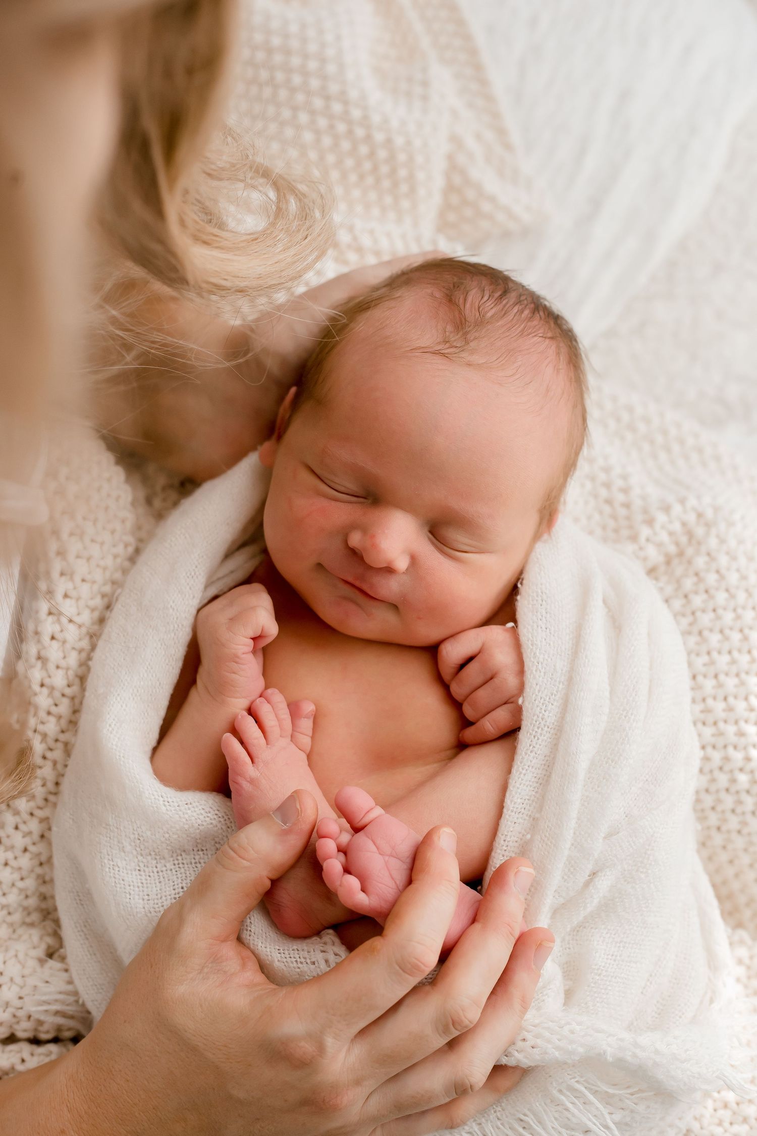 Sleeping newborn baby wrapped in white blanket while hands gently touch tiny feet.