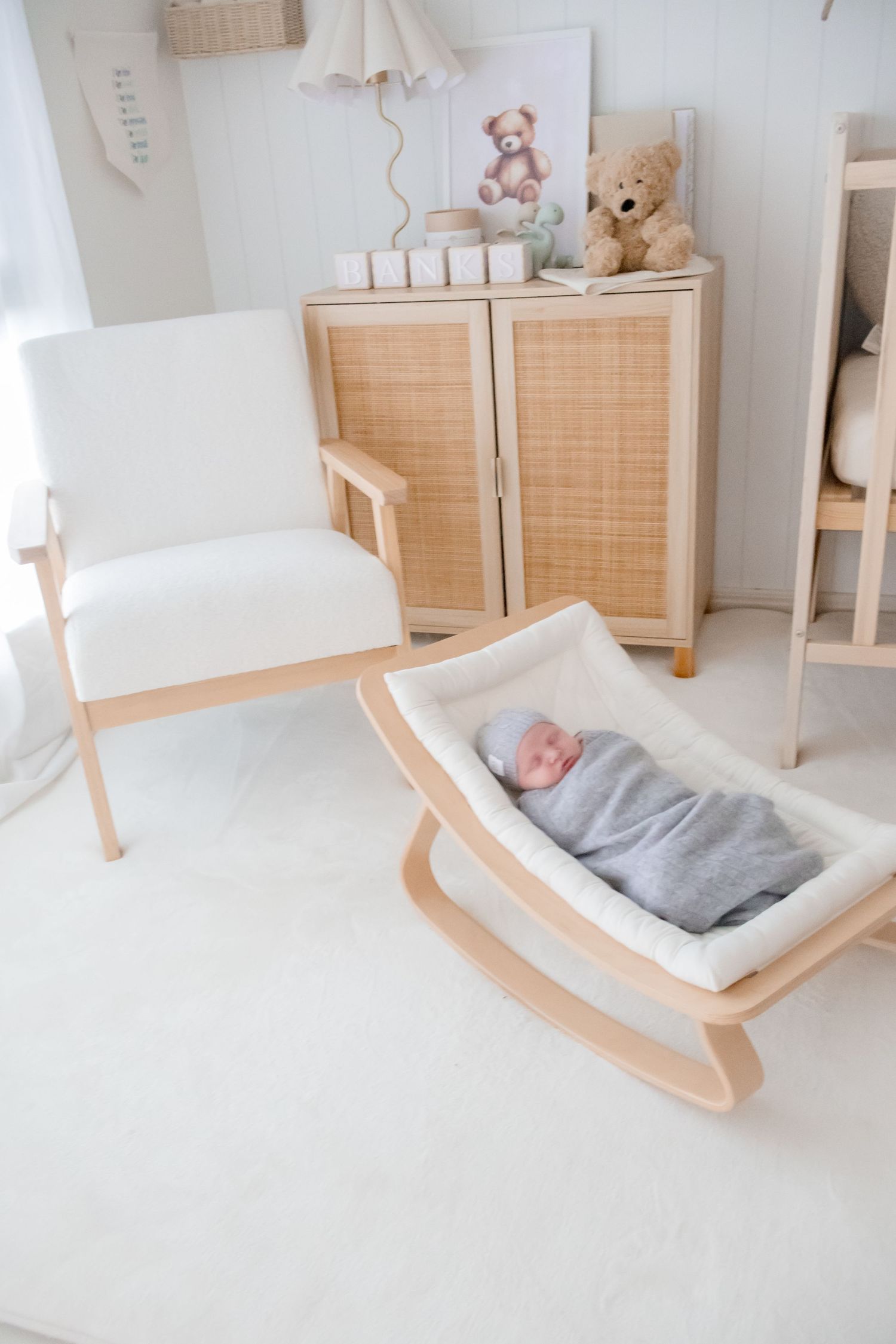 Light and airy nursery with wooden rocking cradle, rattan cabinet, and soft teddy bears in minimalist white decor.