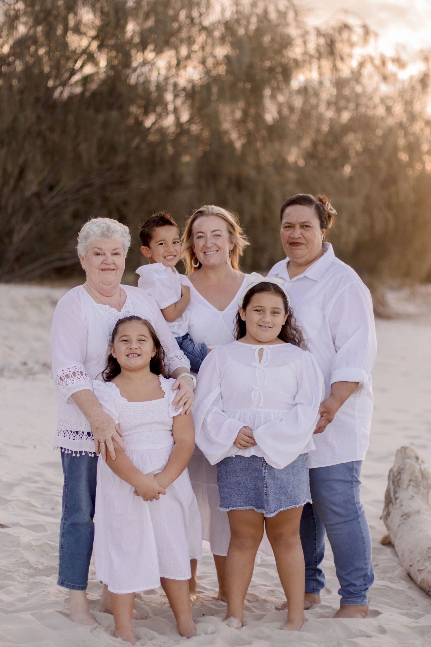 A family wearing white and blue casual outfits poses together on a sunny beach at golden hour.