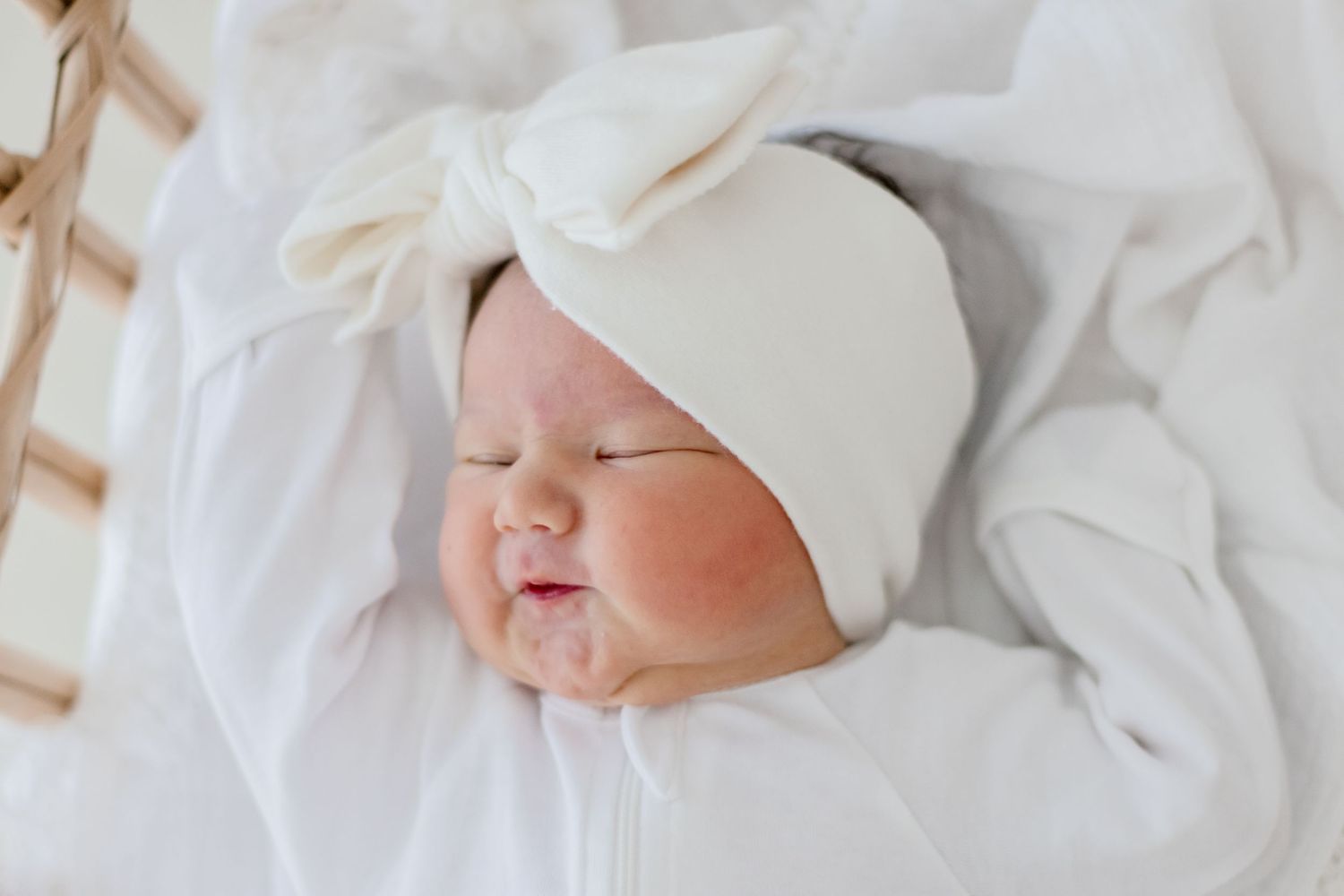 A sleeping baby wearing a white knotted hat rests on soft white bedding.