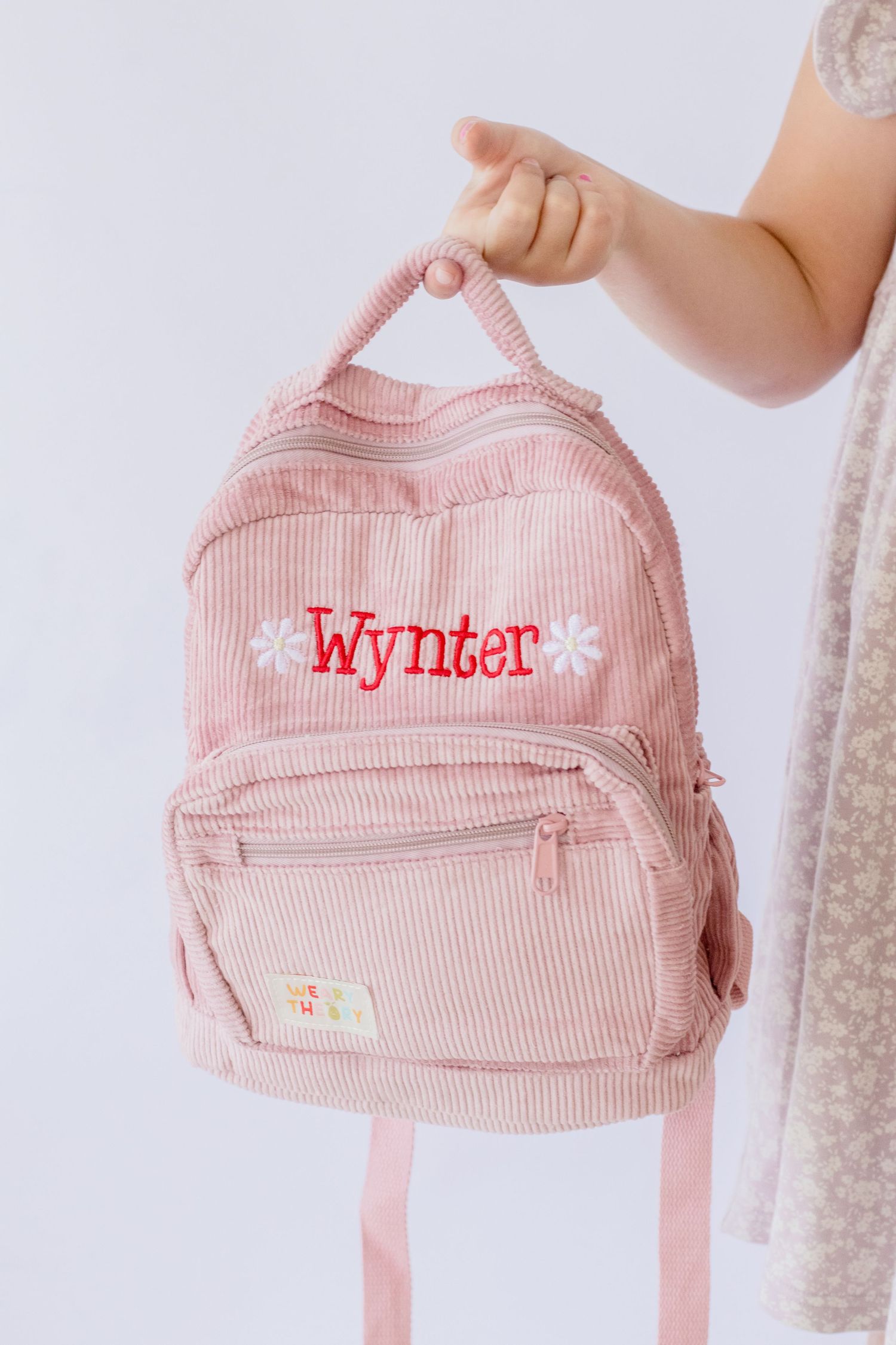 A pink corduroy backpack with 'Wynter' personalized embroidery is held against a white background.