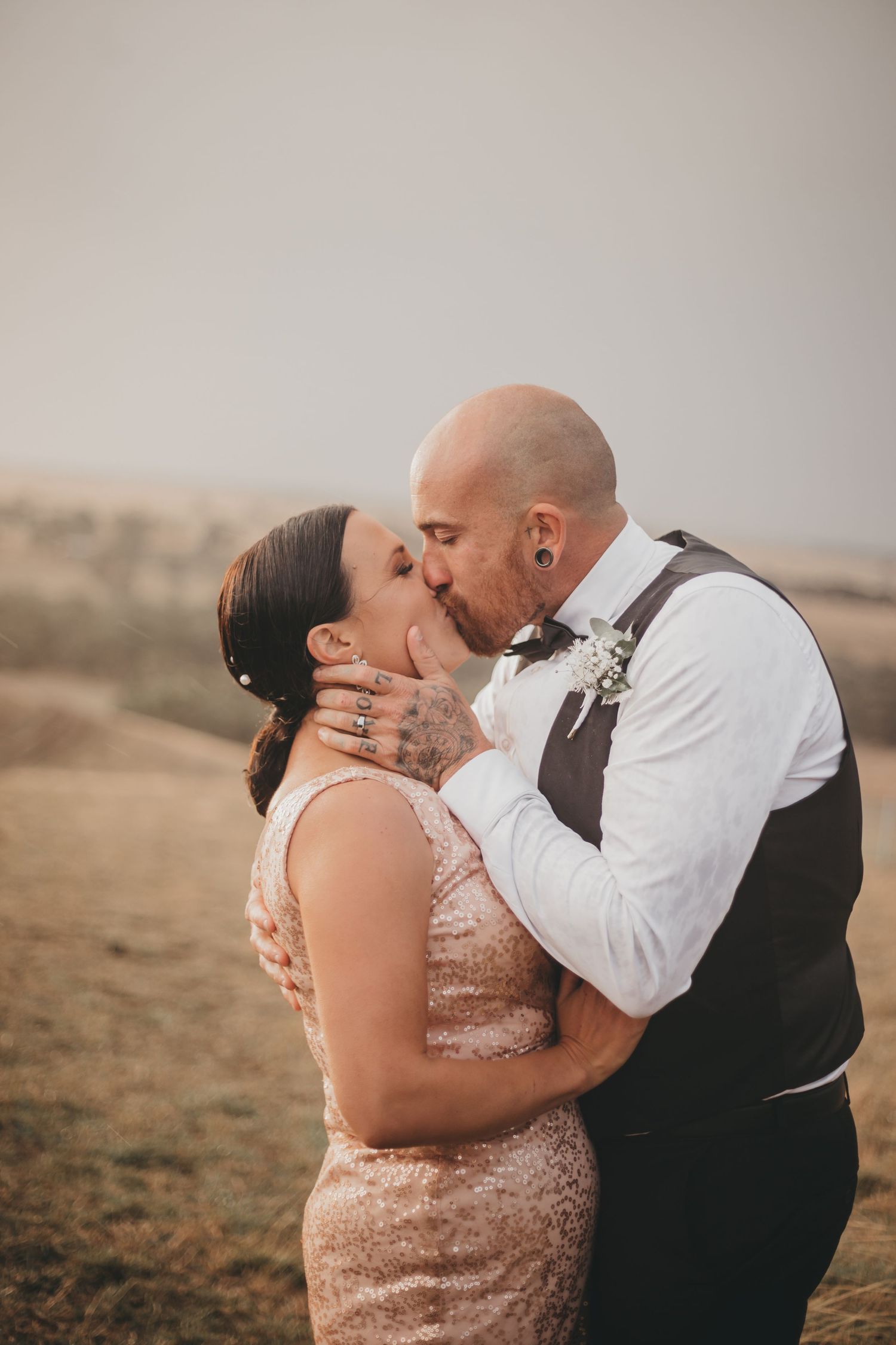 A romantic wedding photo showing a couple kissing outdoors at sunset, with distant hills as the backdrop.