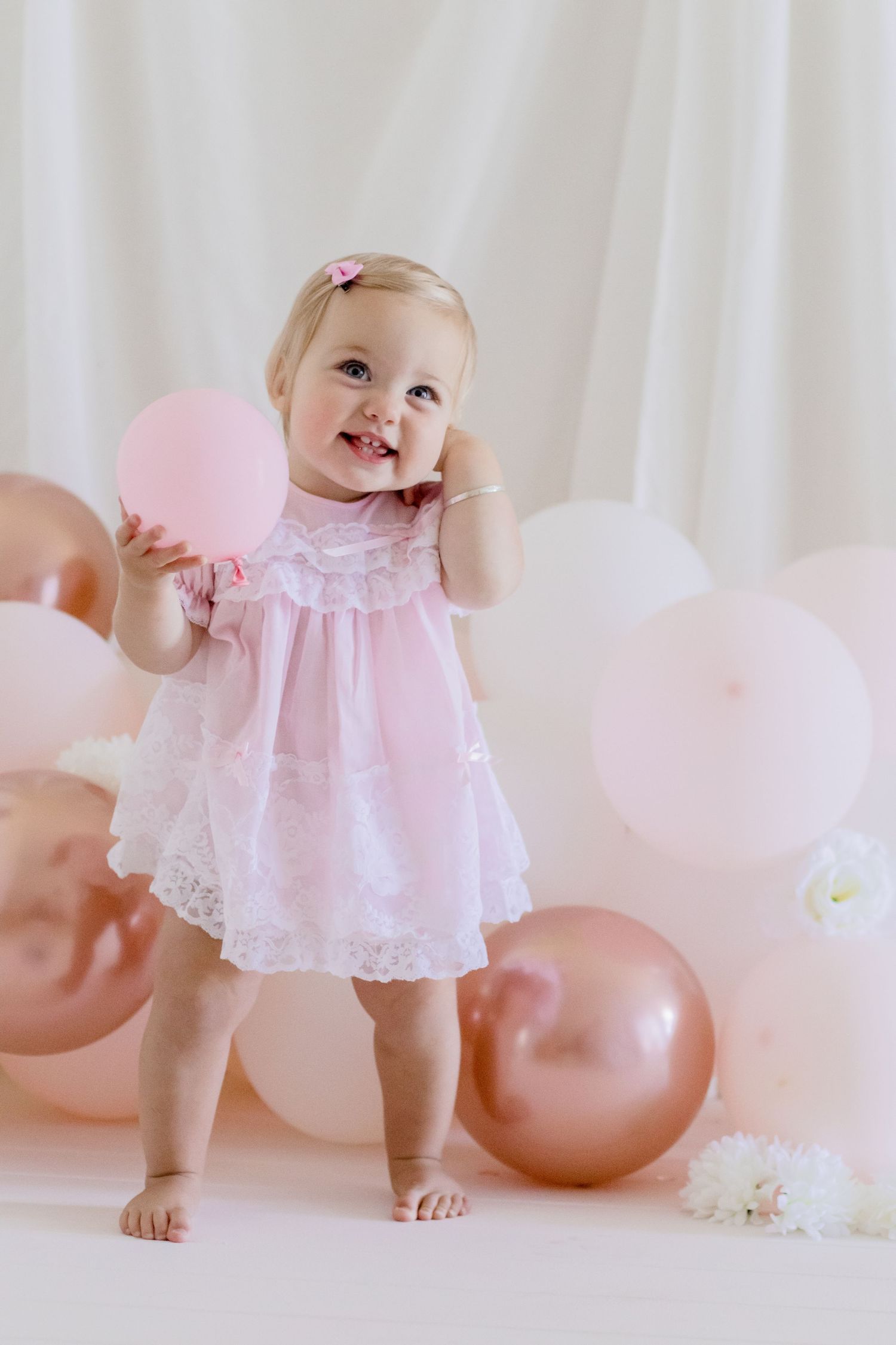Smiling toddler in delicate pink dress plays with balloons against white background.